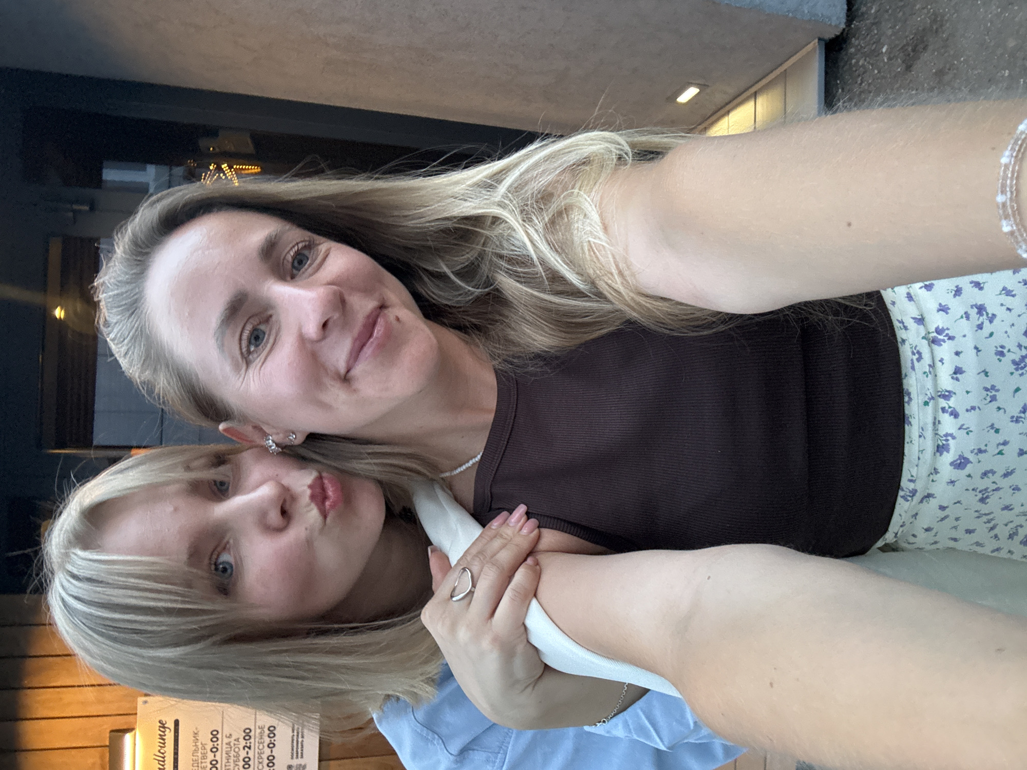 Two women are taking a selfie outdoors in the city of Chaikovsky, Russia. The woman on the left has blonde hair with bangs and is leaning in to kiss the woman on the right, who has long blonde hair and is smiling at the camera. The woman on the left is wearing a light blue shirt, and the woman on the right is wearing a dark brown tank top and a light-colored skirt with a floral pattern. The woman on the left is wearing a silver ring on her right hand, which is resting on the arm of the woman on the right. A sign with Russian text is visible in the background, along with a building facade and a street lamp.