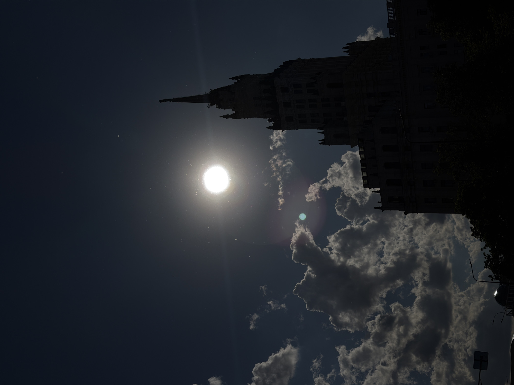 The image presents a dramatic architectural scene, dominated by the silhouette of a tall, ornate building against a bright sky. The structure, strongly resembling one of Moscow's "Seven Sisters" Stalinist skyscrapers with its distinctive spire and intricate gothic-revival details, occupies the right side of the frame, rendered almost entirely in shadow.
In the middle-left, the sun appears as a brilliant, overexposed white disc, surrounded by a strong halo and various lens flares and light streaks, indicating its powerful presence and low angle. Numerous small, bright specks, possibly dust on the lens or sensor, are scattered across the sky, particularly in the vicinity of the sun.
The sky itself is a deep, dark blue-grey, suggesting either early morning, late afternoon, or heavily overcast conditions. Fluffy cumulus clouds are visible, primarily in the lower right and mid-ground; some are brightly illuminated at their edges by the sun, while others remain darker. Dark, indistinct shapes of trees or foliage can be glimpsed on the far right, further silhouetted against the sky.
The composition emphasizes the contrast between the luminous sun and the dark, imposing architecture, creating a powerful and somewhat mysterious atmosphere. No discernible text is visible in the image.