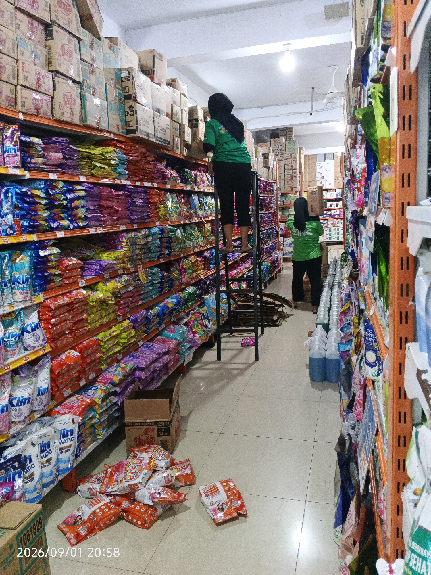 The image depicts the interior of a retail store, likely a minimarket or supermarket, located in Pare-Pare, Indonesia. The store is well-stocked, with shelves lined with various packaged goods, predominantly household cleaning products and detergents, visible on the left side. On the right side, more items are displayed on orange metal shelving units, including bottles and spray containers.
In the foreground, on the floor, are several packages of what appear to be snacks or food items, predominantly in orange packaging. A cardboard box with the number "1260143" printed on it is also on the floor, situated near the shelves.
Two individuals are present in the scene, both wearing green shirts and dark pants. One person is standing on a black metal stepladder, reaching towards the upper shelves, and is partially obscured by the ladder. The second person is in the middle of the aisle, facing away from the camera, carrying a cardboard box on their head. This individual appears to be in the process of stocking or moving inventory.
The environment is brightly lit with fluorescent lighting on the ceiling. The floor is tiled with a light-colored, possibly ceramic, material. A ceiling fan is visible, suggesting the store is air-conditioned or at least has air circulation. The overall impression is one of an active retail environment where restocking and organization are in progress. The date stamp at the bottom left corner indicates "2026/09/01 20:58".