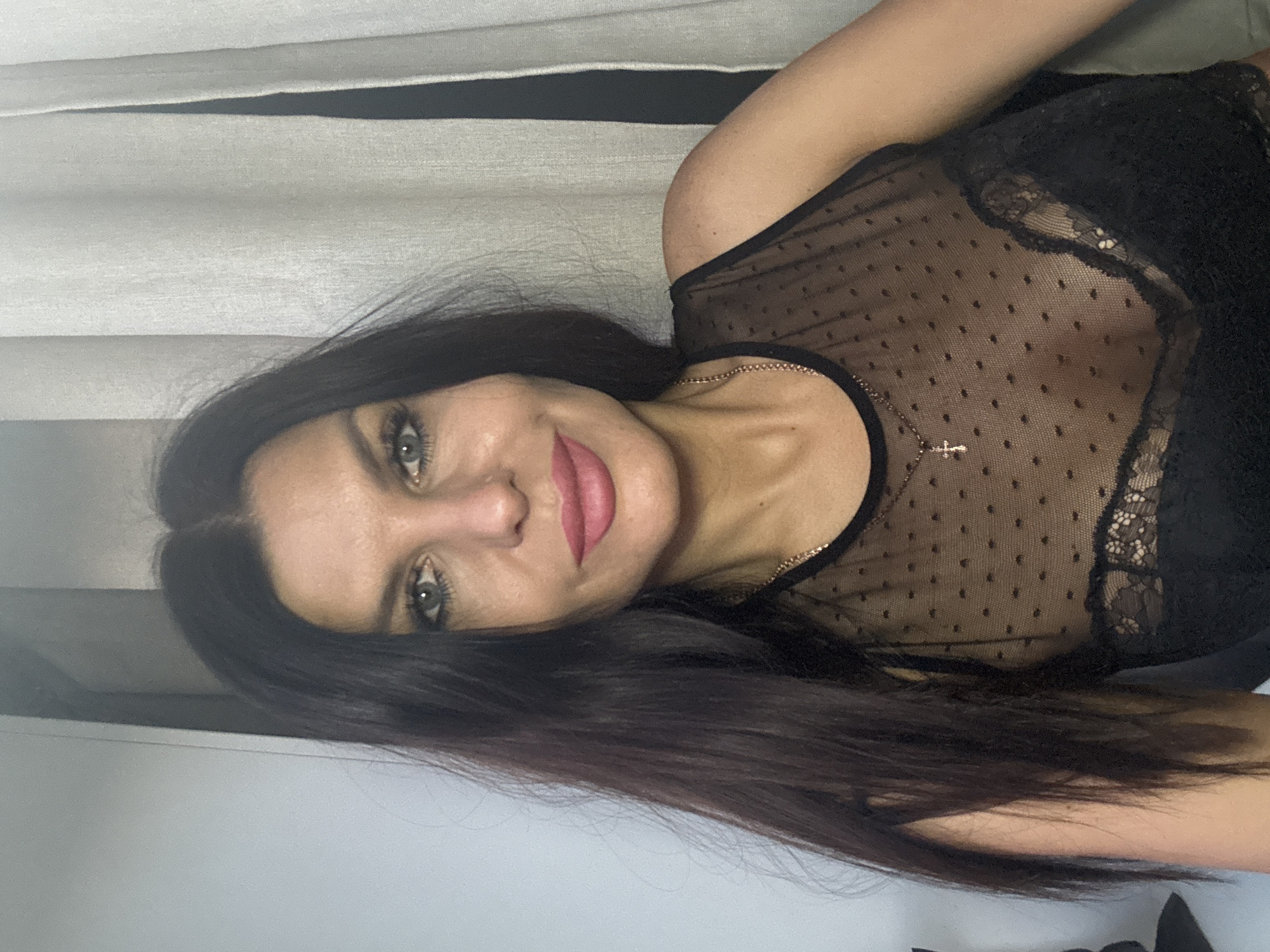 A woman with long, dark hair and light-colored eyes is featured in this horizontally oriented portrait. She has prominent makeup, including dark eyeliner and pink lipstick, and looks directly at the camera with a gentle smile, her head slightly tilted. She is wearing a black sheer top adorned with small polka dots and lace detailing along the neckline and shoulders. A delicate gold chain with a small cross pendant rests on her collarbone. Her dark hair frames her face and flows over her shoulders and arm. The background consists of light-colored fabric with horizontal dark stripes, possibly indicating blinds or curtains in an indoor setting. There is no visible text. The image was taken in Chaikovsky, Russia.