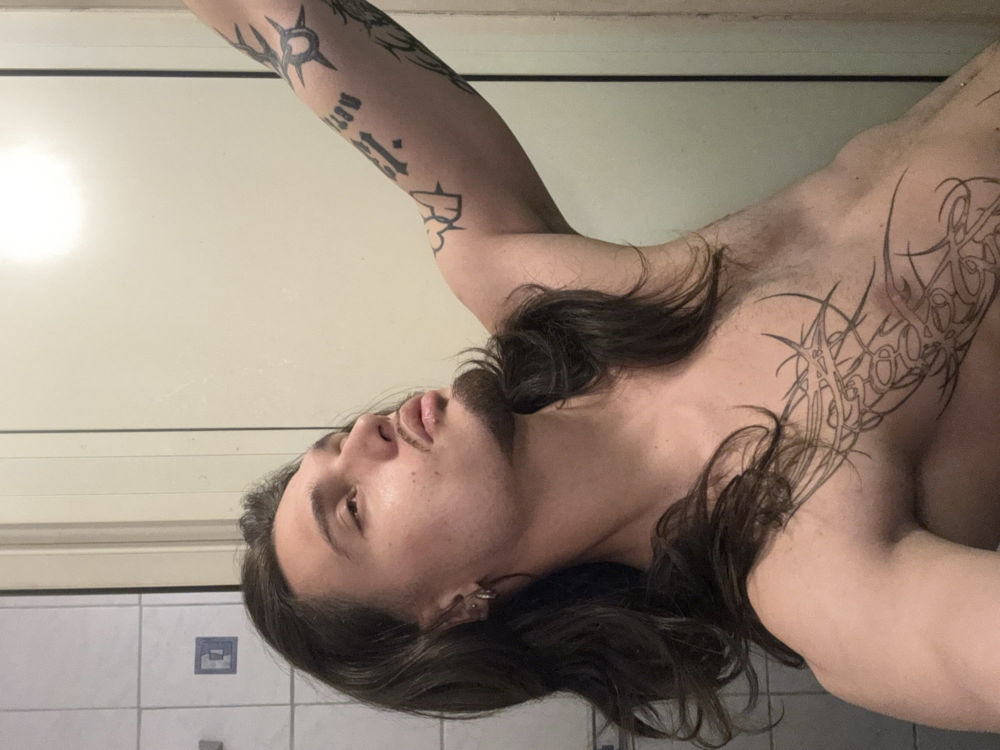 The image, rotated 90 degrees clockwise, captures a close-up of a person with long, dark, wavy hair and extensive tattoos, likely indoors in Sofia, Bulgaria.
**Subjects:** The central subject is a young adult, possibly male, with a light complexion, a neatly trimmed mustache, and a subtle beard. Their dark hair cascades over their left shoulder and chest. They are shirtless, prominently displaying several tattoos. A small, silver-colored ring or hoop is visible in their left earlobe.
**Objects:** The person is adorned with numerous tattoos. On their right arm (appearing on the left side of the rotated image), there are several distinct designs, including a starburst or sun-like symbol and what appear to be stylized script or numbers, with "13" being partially discernible among other characters. Their left chest and shoulder feature a large, intricate tribal or abstract design with swirling lines and sharp, pointed elements. More script-like tattoos are visible on their left arm.
**Scene/Setting:** The setting appears to be an indoor space, possibly a bathroom, indicated by the white rectangular tiles with dark grout lines visible in the lower part of the image (left side in the original orientation). Above the tiles is a light-colored wall, possibly painted off-white or beige, with a dark horizontal line, suggesting a trim or a transition between wall sections. A small blue and white patterned tile or decorative element is visible on the tiled wall.
**Activity or event taking place:** The person is captured in a posed shot, looking upwards with a slightly open mouth, giving a contemplative or neutral expression. Their right arm is raised above their head. The scene suggests a self-portrait or a deliberately composed photograph.
**Notable details:** The dominant lighting is artificial, casting highlights on the person's skin and hair. There are no strong emotional cues, and the time of day is not discernible from the indoor lighting. The visible tattoos contribute significantly to the person's aesthetic.