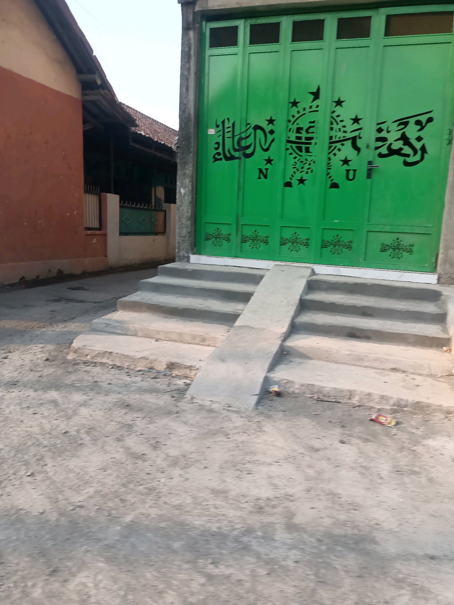 The image shows a street scene in Pecabean, Indonesia, with a focus on a bright green folding gate adorned with black Arabic calligraphy and star motifs. The gate is part of a building with a rough concrete facade to its right. In front of the gate, there are several concrete steps leading up to it, with a concrete ramp on the left side of the steps. The ground in front of the steps is unpaved and appears to be dirt or gravel. To the left of the steps and gate, there is a building with an orange-brown wall and a sloped, tiled roof. Further back, between the two buildings, a glimpse of a fence and another structure with a tiled roof can be seen. The sky is visible above, appearing pale and possibly overcast. The scene suggests an outdoor, potentially residential or commercial, setting in a village or town.