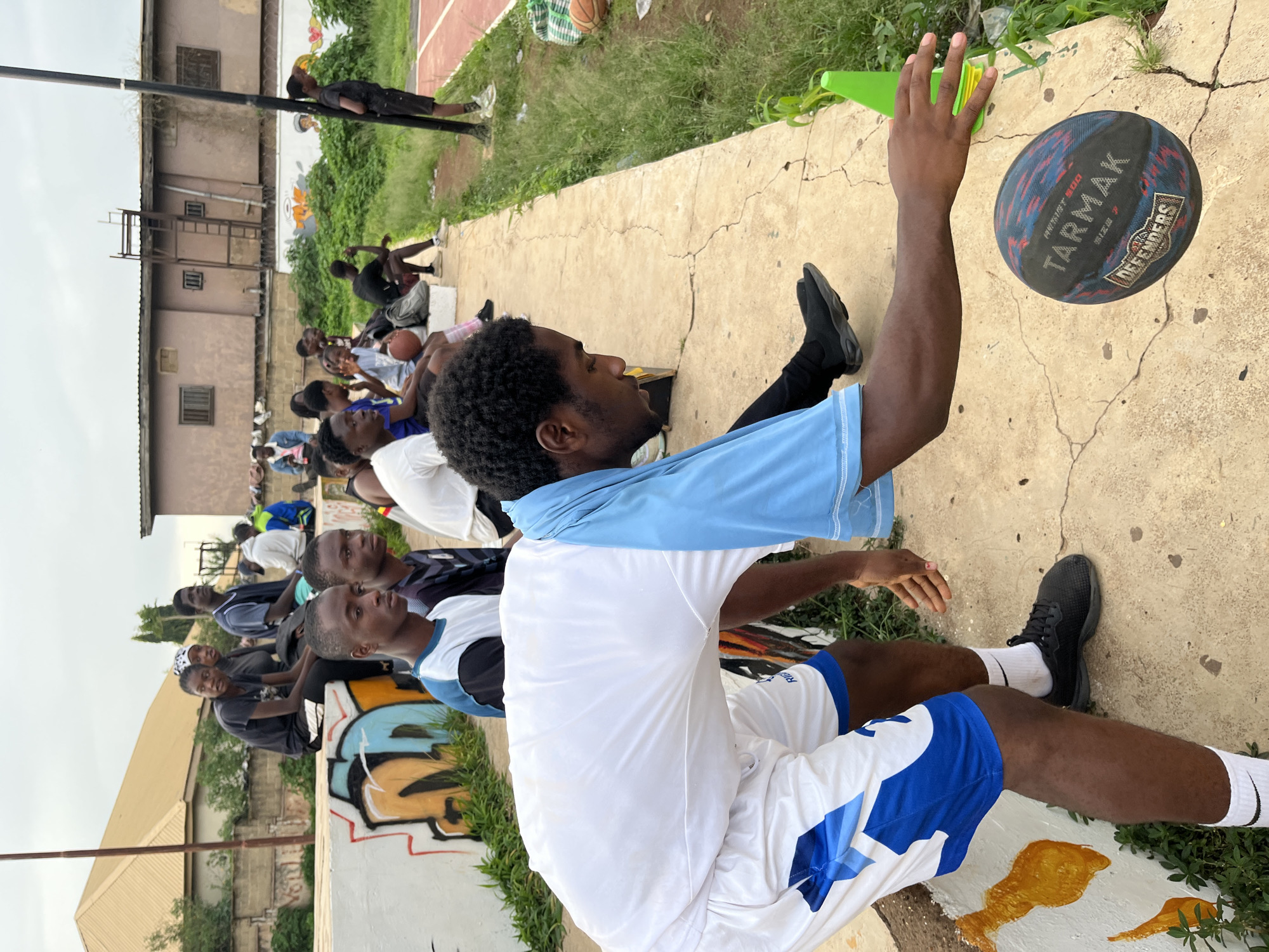 The image captures an outdoor basketball training session in Gura Top, Nigeria. In the foreground, a young Black male athlete, wearing a white shirt with light blue sleeves and white shorts with blue accents, is positioned in a low stance, dribbling a Tarmak basketball. His right arm is extended towards the ball, with his hand positioned above it. Nearby, a green traffic cone is visible. In the middle ground, a group of young Black individuals are seated on a concrete bench, some wearing athletic attire, observing the activity. Behind them, a white concrete wall is adorned with graffiti featuring abstract shapes and colors. Further in the background, a two-story building with a pale brown facade stands, with a metal ladder attached to its side. Overgrown green vegetation flanks the concrete bench and stretches towards the distance. The sky is overcast, indicating diffused lighting conditions. The overall scene depicts a casual sports gathering in a semi-urban environment.