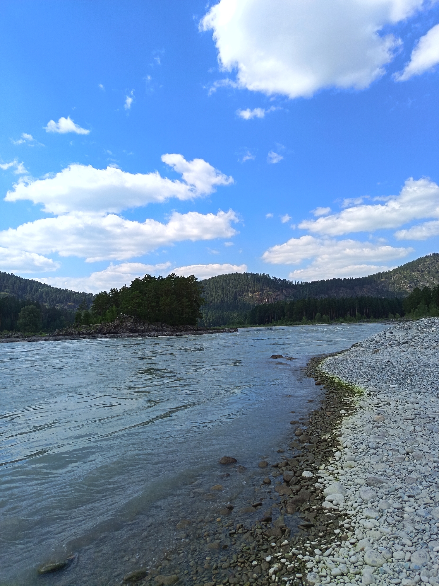 The image depicts a wide, rocky river flowing through a landscape dominated by forested mountains under a blue sky with scattered white clouds. The river occupies the left and central portions of the frame, its surface rippled and showing a bluish-gray hue. The right bank is densely covered with smooth, rounded pebbles of various sizes, creating a distinct texture. In the middle distance, a small, tree-covered island is visible, surrounded by rocks. The background features rolling hills and mountains covered in dark green coniferous forests. The sky is a vibrant blue, with fluffy white clouds casting shadows on the landscape. No people or structures are visible in the image. The scene is one of natural wilderness, likely a remote area in Russia, given the mention of "Нижнекаянча".