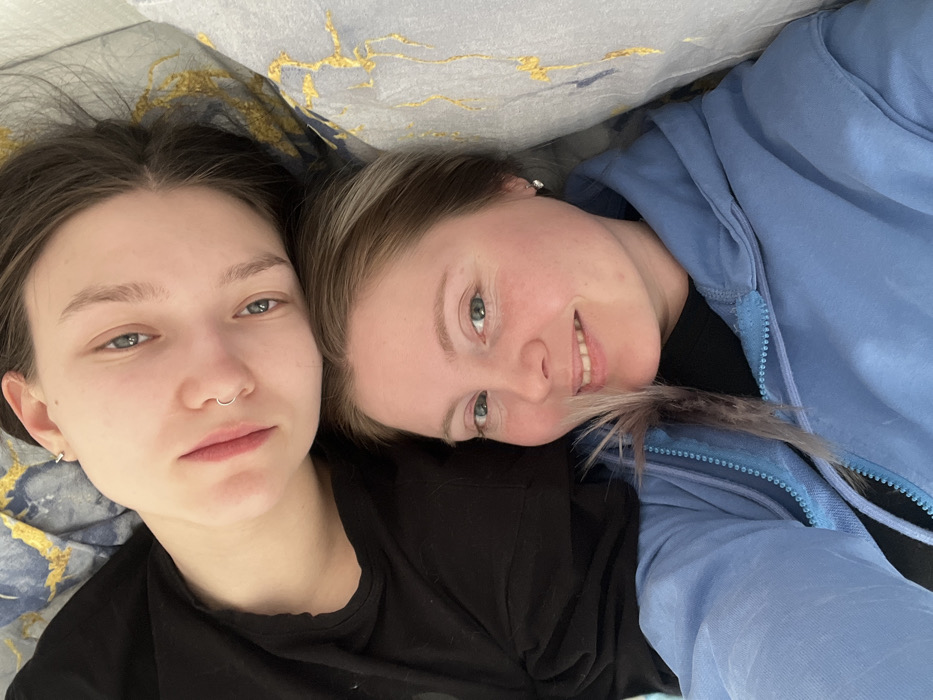 The image depicts two females positioned closely together in what appears to be an indoor residential setting. The individual occupying the upper portion of the frame has dark hair, light blue eyes, a septum piercing, and a small hoop earring in her left ear, wearing a black top. Her head is resting near the second individual. The individual in the lower portion of the frame has light brown hair with blonde highlights, light blue-green eyes, and a small stud earring in her right ear, wearing a light blue zippered hoodie over a black top. A lock of her hair near her face shows a light purple or grey tint. She is smiling towards the camera, while the upper individual maintains a neutral expression. The background consists of bedding or pillows; one features a dark blue and gold abstract pattern, and the other is light grey. The specific location within the city of Chaikovsky, Russia, is not discernible from the visual evidence.