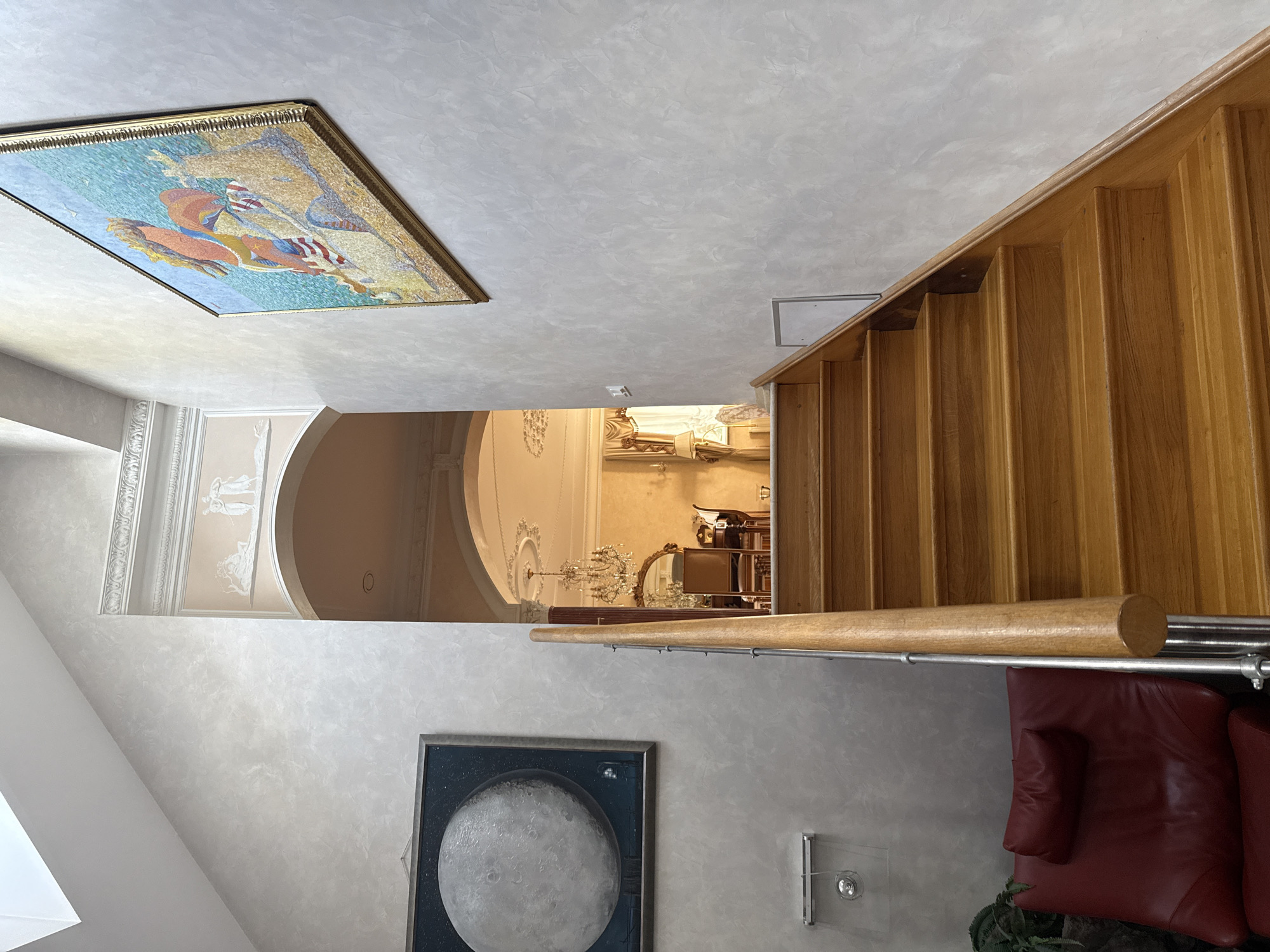 This image captures a well-lit interior hallway featuring a prominent wooden staircase. The hallway walls are textured, with one wall displaying a framed painting of the moon and the other a large, framed mosaic artwork of a figure with a horse against a blue background. An arched doorway leads into another room, revealing a chandelier, ornate ceiling decor, and upholstered furniture. A red leather armchair is situated at the base of the stairs. The scene is set in the city of Yukki, Russia.