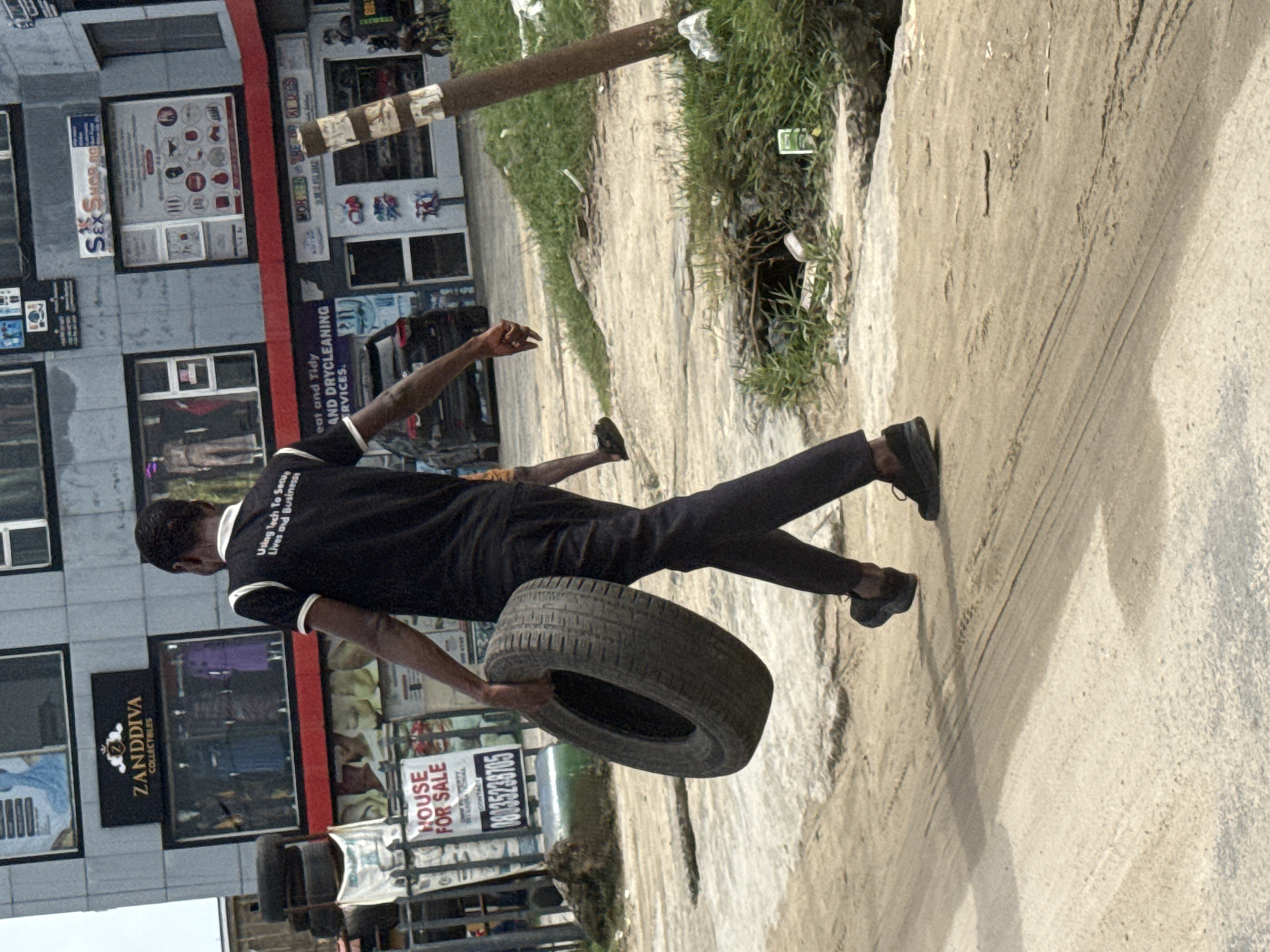 The image captures a man in motion on a street in Lekki, Nigeria. He is carrying a tire. He is wearing black trousers, a black t-shirt with white detailing, and black shoes. The background includes a building with visible businesses, including ZANDDIVA COLLECTIBLES and a dry cleaning service. A "HOUSE FOR SALE" sign is also visible. The street surface is rough, showing tire tracks. There is some vegetation and a utility pole to the right of the man. The image is rotated.