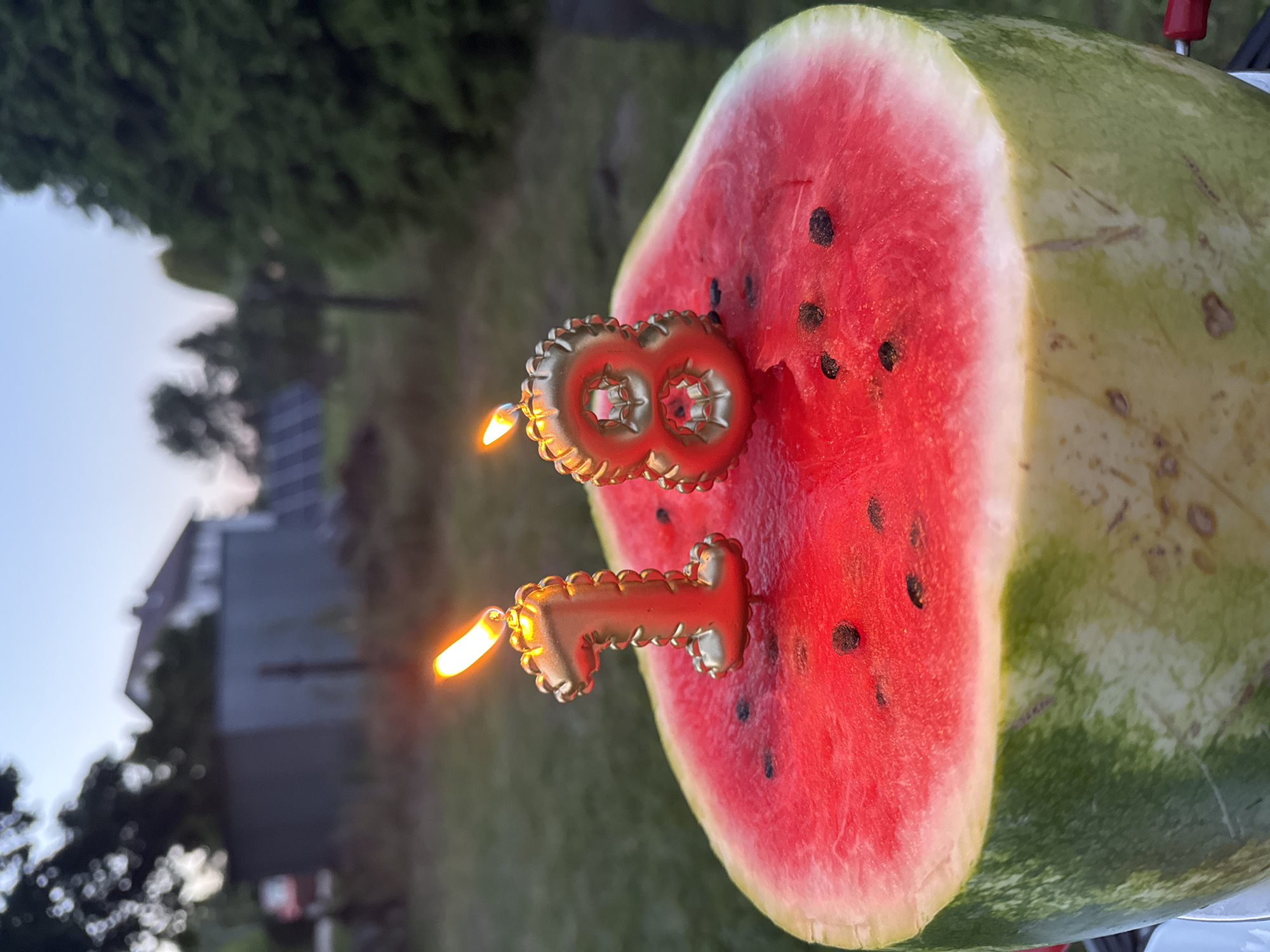 A close-up shot focuses on a large slice of watermelon, the bright red flesh speckled with black seeds, topped with two gold-colored, lit birthday candles shaped like the numbers "1" and "8". The candles are slightly tilted, their flames flickering upwards, casting a warm glow on the fruit. The watermelon is positioned outdoors, with a blurry background suggesting a yard. In the distance, a house with a dark roof is visible, along with trees and a fence. The sky is a pale blue, indicating it might be dusk or dawn. The overall scene conveys a celebratory atmosphere, likely marking an 18th birthday. The specific location in Katowice, Poland, cannot be determined from the visual information provided in the image.