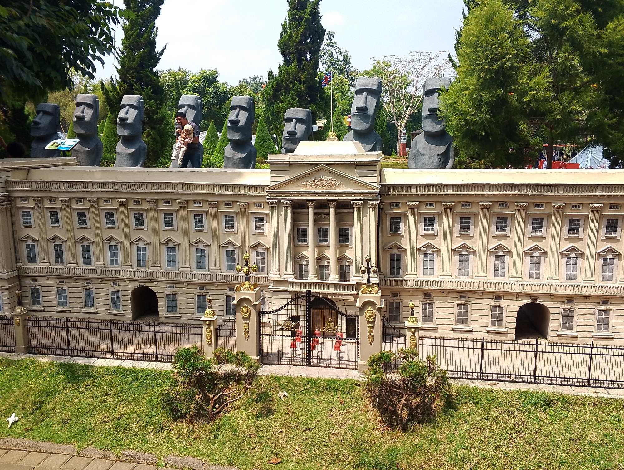The image depicts a miniaturized replica of a royal palace, likely Buckingham Palace, situated in the city of Megamendung, Indonesia, within the Taman Safari area. In front of the miniature palace, a row of Moai statues from Easter Island are prominently displayed. A man is seen holding a child, both dressed casually, standing next to one of the Moai statues. The palace replica features a classical architectural style with numerous windows, columns, and decorative elements. A detailed wrought-iron gate guards the entrance, with four small figures in red uniforms standing in formation behind it. Lush green grass covers the foreground, with small bushes and trees dotting the landscape, suggesting an outdoor recreational park setting. The sky is clear and bright, indicating a sunny day.