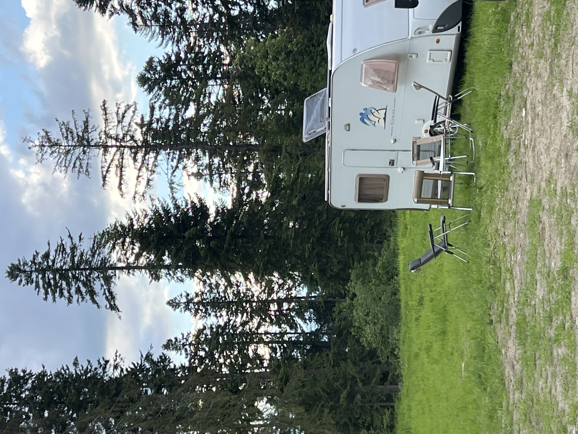 An outdoor scene depicts a white caravan parked in a grassy area with tall evergreen trees in the background. A few folding chairs are set up in front of the caravan. The sky is partly cloudy. The caravan has a small logo with two figures and a mountain or wave graphic. The location is identified as Unknown, Unknown.