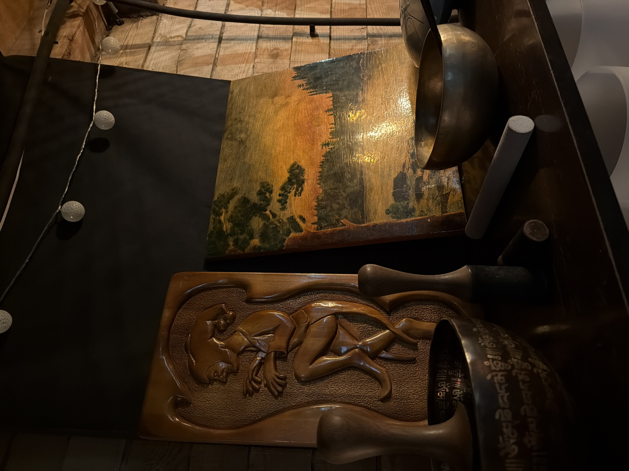 The image captures a dimly lit corner, possibly inside a shop or gallery. A wooden carving featuring a human figure in a reclining position dominates the lower section. Above it sits a painted landscape, depicting trees and a forest-like setting with a predominantly yellow and green color palette. Several metal bowls or gongs are positioned to the side, obscuring parts of the artwork. A string of decorative lights with spherical bulbs runs along the upper left side of the frame. The background reveals wooden planks and dark surfaces. There is no information about Krasnoyarsk, Russia.