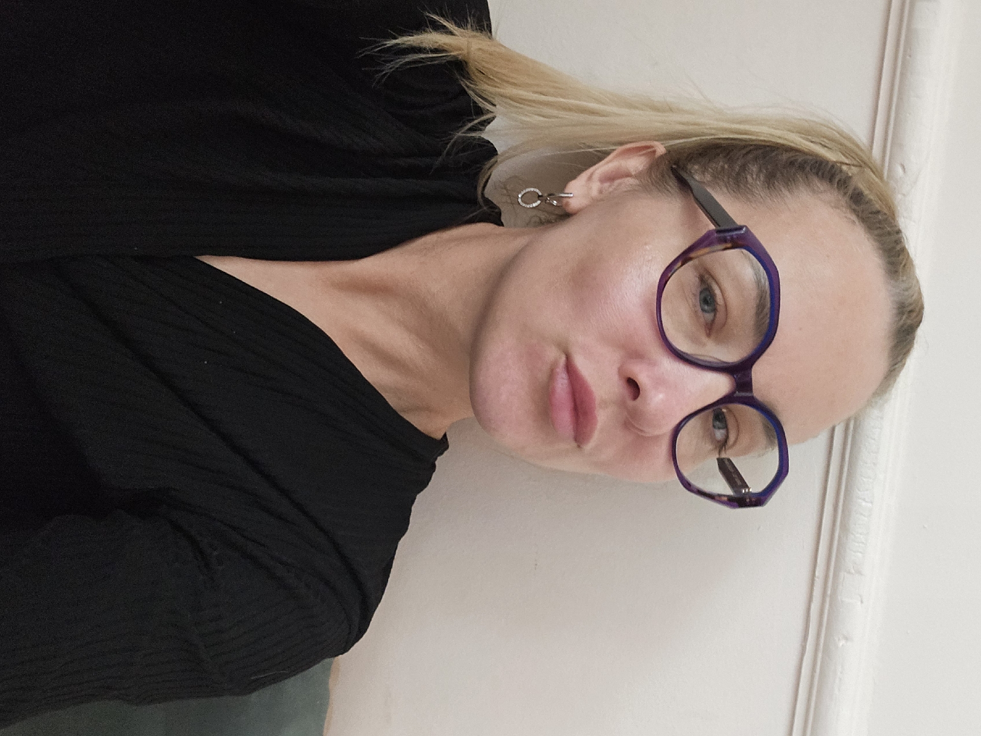 A woman with light blond hair and wearing purple-rimmed glasses is positioned against a white wall. She is wearing a black top. She has light skin, and her hair is pulled back. She is wearing small, circular earrings. The image is taken at an angle. There are no indications of Sochi, Russia.