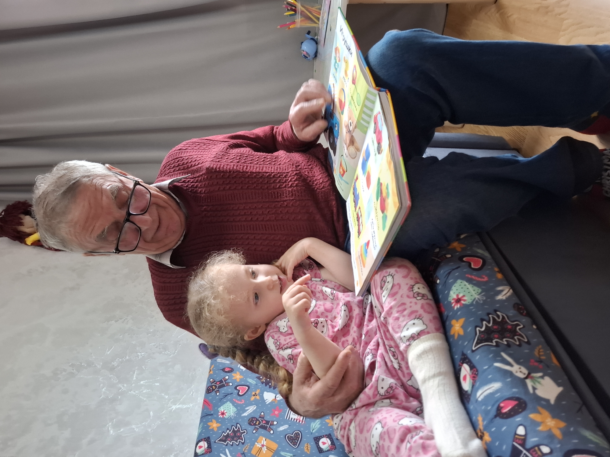 An older man wearing glasses and a maroon sweater is reading a picture book to a young girl in pink Hello Kitty pajamas. The girl is sitting in the man's lap, with her right hand near her mouth. They are sitting on a patterned pillow. In the background are a gray curtain, wooden desk, and a container of colored pencils. The location is Chaikovsky, Russia.