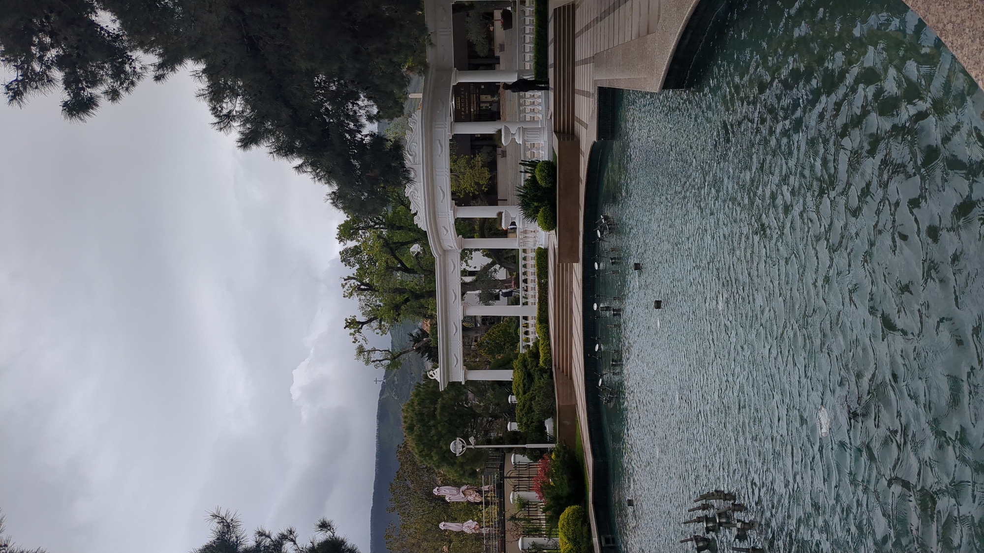 The image shows a scene in Gelendzhik, Russia, featuring a park or garden area with a large pool or fountain. The water is somewhat reflective, showing ripples and disturbances. A white colonnade or gazebo-like structure stands adjacent to the water's edge. The architecture is classical, with columns and decorative elements. A person in dark clothing is visible near the structure. The background includes trees, mountains shrouded in cloud or mist, and an overcast sky. Streetlamps, landscaping, and seating areas are present, suggesting a well-maintained public space.
