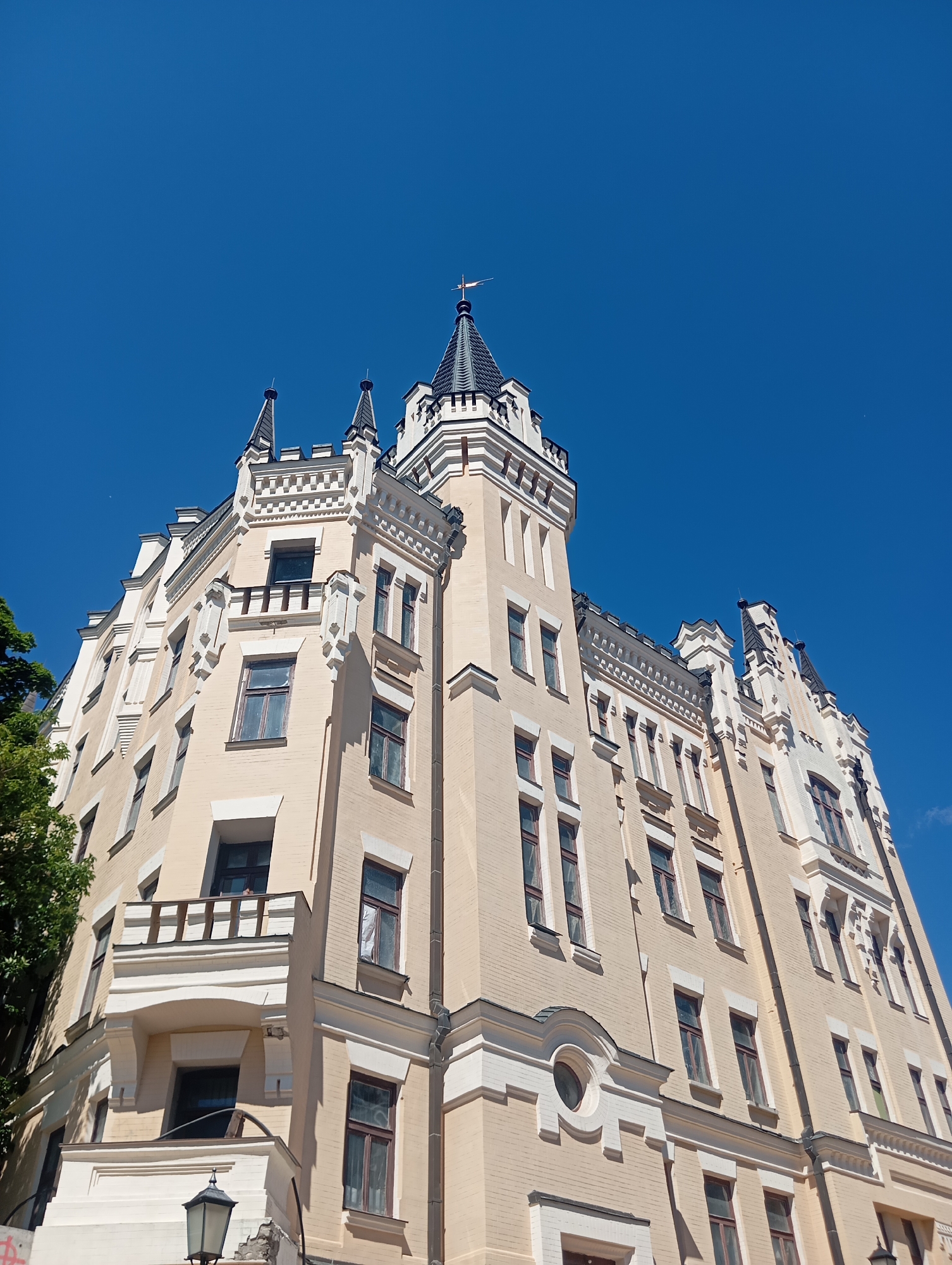 This is a low-angle, full shot of a pale yellow building with gothic architectural elements, set against a clear blue sky. The building features multiple towers with pointed, dark roofs, decorative white trim, and numerous windows. Some of the windows have dark frames, while others appear to be boarded up or covered with white material. A small balcony with a wooden railing is visible on one side of the building, and a street lamp is present in the lower left corner. A tree with green foliage is partially visible on the left edge of the image. The building is located in Kyiv, Ukraine.