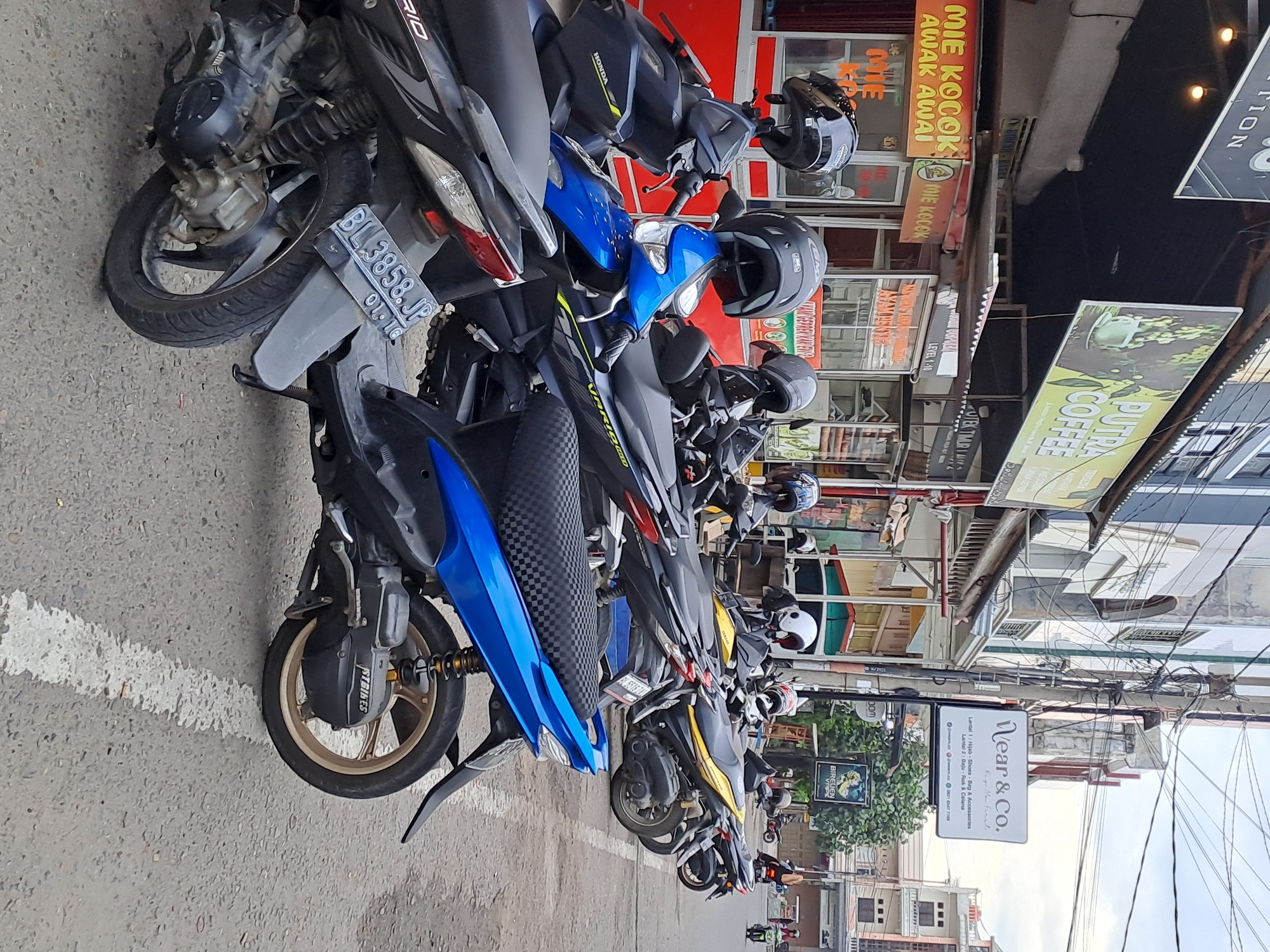The image depicts a street scene in Bandarbireuen, Indonesia, with a row of parked motorcycles on the right side of the road. Several shops are visible in the background, including one with a sign that reads "PUTRA COFFEE" and another advertising "MIE KOCONK AWAK AWAI". A sign for "Wear & Co." which sells Hijab, Shoes, Bags & Accessories, and Clothes is also present. The sky is overcast. One motorcycle in the foreground is blue and black, while others are black, dark blue, and yellow. Helmets are placed on top of many of the motorcycles. The street is paved and marked with a white line.