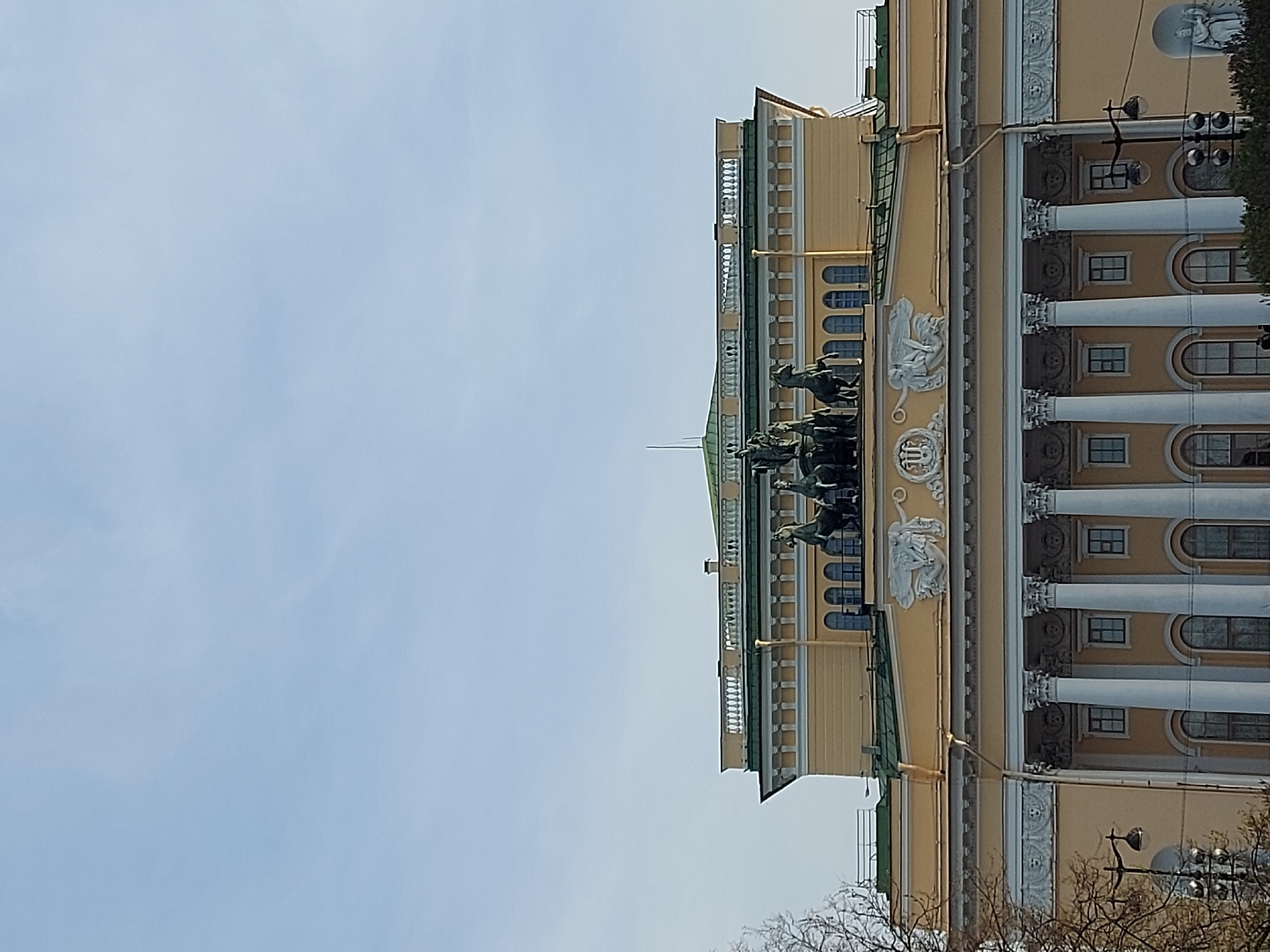 Here is a detailed description of the image:
The image shows a building in Saint Petersburg, Russia. It appears to be the General Staff Building, located on Palace Square. The angle of the photo shows the building from the side, with a large, open sky dominating the left side of the frame.
The building is a pale yellow color and features a classical architectural style, with prominent columns and decorative elements. A notable detail is the sculptural group of a chariot pulled by horses, positioned on top of the building. The building has multiple levels with arched windows and ornamental details between the columns.
The sky is a light blue color and appears clear.
The image is a static outdoor shot taken during daylight.
