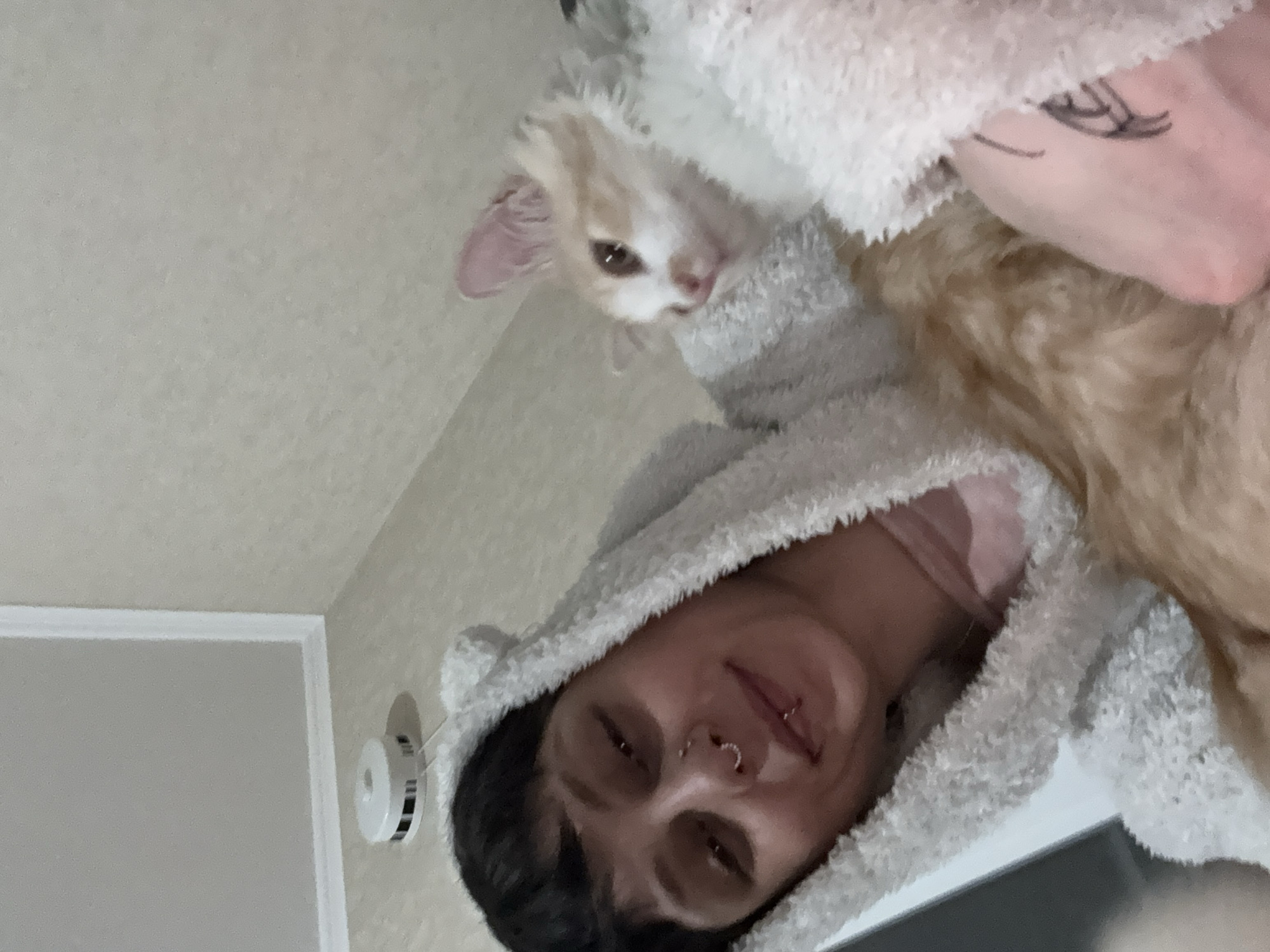 An indoor scene depicts an individual and a cat in a close-up, selfie-style photograph. The individual, positioned on the left, has short, dark hair and is wearing a light-colored, fuzzy hooded garment with ear-like protrusions. They present a subtle smile, featuring a silver septum piercing and two silver rings on the lower lip. A pink garment is visible beneath the fuzzy exterior. A partially obscured dark tattoo or drawing is present on the individual's left forearm or hand. A light orange and white cat is situated on the right, partially enveloped by the individual and the fuzzy garment, with its head turned to the right and pink ears visible. In the upper left background, a white, circular smoke detector with black stripes is mounted on a light-colored wall at its junction with the ceiling, where white molding is also visible. The walls and ceiling are a uniform, light, neutral color. The specific location within the city of Saint Petersburg, Russia, cannot be determined from the visual information provided.