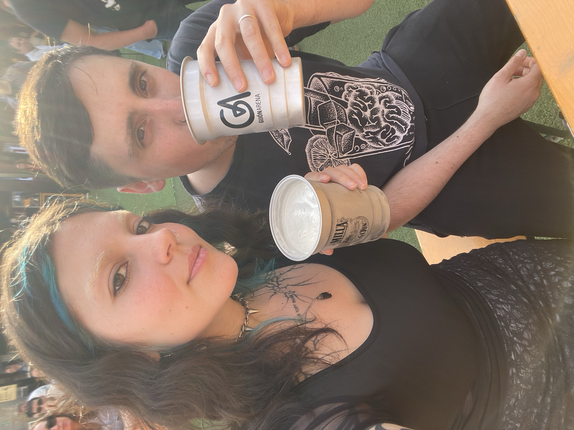 A couple poses for a selfie outdoors in Gijón/Xixón, Spain. The woman, in the foreground, has dark hair with blue streaks, a nose ring, and a spiked choker necklace. She wears a black top over a patterned mesh shirt. A tattoo is visible on her collarbone. The man, slightly behind, holds a white paper cup with the inscription "GUONARENA" and a black logo, appearing to drink from it. He also wears a black t-shirt with a white graphic print and a wedding ring on his finger. Both are holding cups of a light-colored beverage. They appear to be near a wooden table, with an artificial green surface visible in the background, along with glimpses of other people and objects. The sunlight is bright, causing flares and shadows.