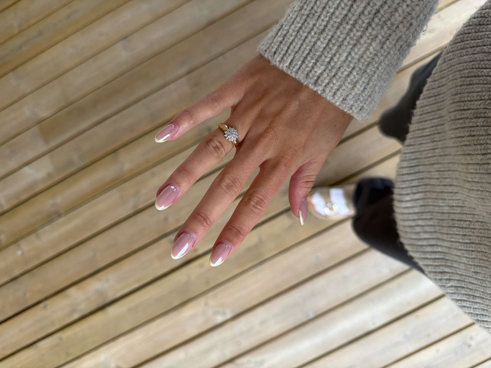 An over-the-shoulder shot from a first-person perspective shows a woman's left hand with a diamond ring on her ring finger and manicured nails with a French tip. The hand is positioned over a wooden deck. The woman is wearing a cream-colored knit sweater and dark pants, with one foot, wearing a sandal, visible at the bottom of the frame. The background is a wooden deck. The scene is not in the city of Harstad, Norway.