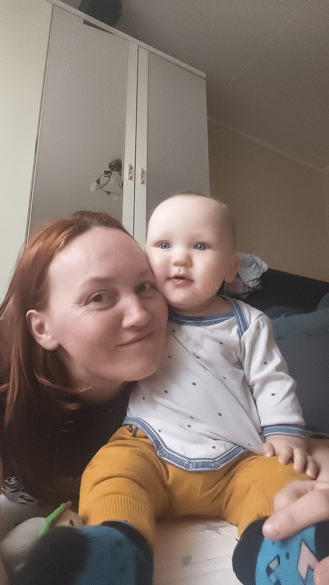 This intimate indoor portrait features a woman and a baby, likely a mother and child, posing closely together. The woman, positioned on the left, has reddish-brown hair and a gentle smile, with subtle lines around her eyes indicating warmth. Her head is slightly tilted towards the baby.
The baby, situated on the right, has striking light blue eyes and looks directly at the viewer with a slightly open mouth, conveying curiosity. The baby is wearing a white long-sleeved onesie with small dark star patterns and light blue trim, mustard yellow pants, and dark-colored socks with light blue and green designs.
They are seated on a light-colored soft surface, possibly a mattress or blanket. The background reveals details of a home environment. To the left, a tall white wardrobe or cabinet with gold-colored handles is visible; one door appears to be mirrored, reflecting a ceiling light fixture with exposed bulbs. To the right, a dark-colored sofa or bed is partially seen against a plain, light-colored wall. A small toy with green and white details is partially visible in the bottom left corner.
The image captures a tender moment between the subjects, filled with affection. The lighting suggests a daytime setting indoors, possibly supplemented by artificial light. There is no visible text.