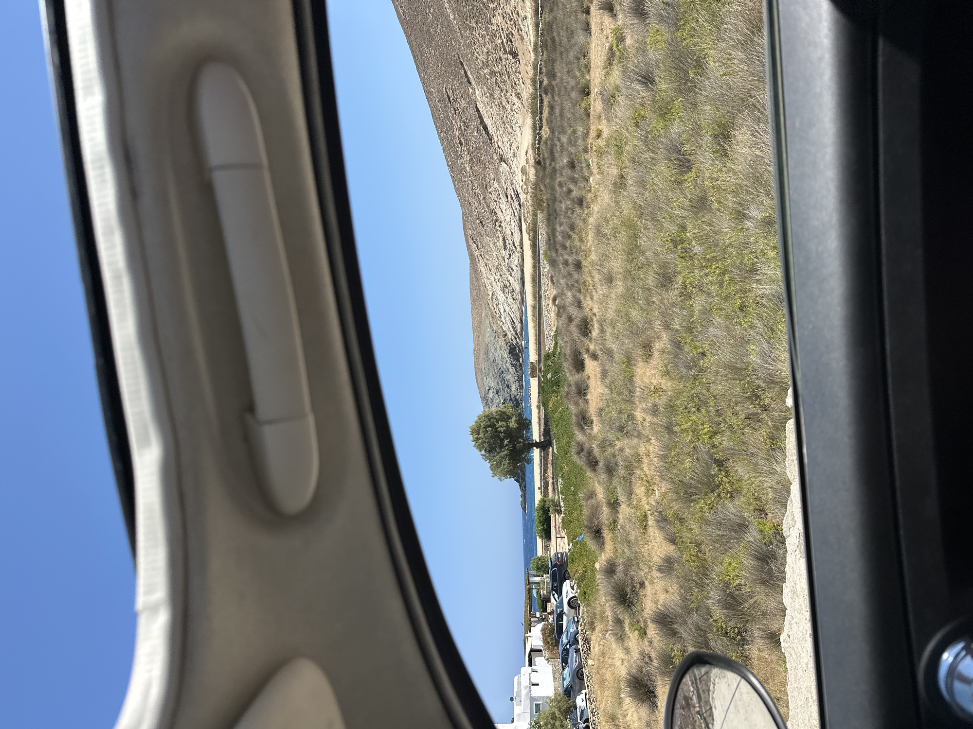 The image, taken from inside a vehicle, looks out onto a sun-drenched landscape in Ano Mera, Greece. The foreground shows a portion of the car's interior, including a black dashboard and a side-view mirror reflecting a dry, grassy terrain. Beyond the car, a parking lot filled with several cars is visible to the left, adjacent to a white building. Further in the distance, a beach meets the calm blue sea. A lone tree stands on the edge of the beach, and rolling, arid hills form the backdrop under a clear blue sky. The scene is characteristic of a coastal area with dry vegetation, common in the Mediterranean climate of Greece.