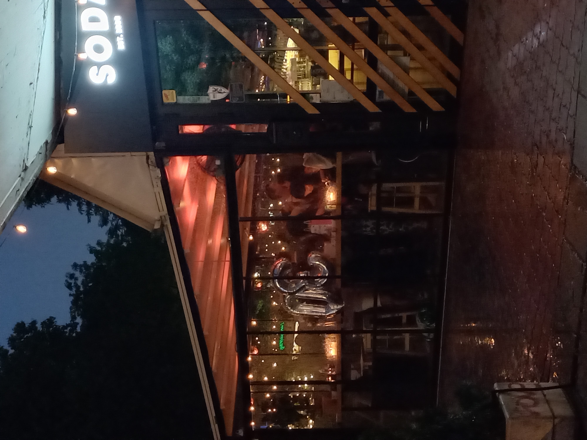 The image captures the exterior of a building at night, likely a restaurant or cafe, in Sofia, Bulgaria. The sky is dark blue, and artificial lights illuminate the scene. The ground in the foreground appears wet and reflective, suggesting recent or ongoing rain, possibly cobblestone paving.
The building features a dark facade with a prominent, illuminated sign on the upper left side that reads "SODA" in large, glowing white letters, with "EST. 2020" inscribed below it. Below the sign, there is a large glass window or wall that offers a glimpse into the interior, which is also dimly lit and shows reflections of the exterior.
The upper part of the building's facade is distinctive, featuring diagonal wooden or metal slats that add a modern design element. To the left of the main entrance, an awning or retractable cover extends outwards, beneath which small string lights are visible, casting a warm glow.
Through the glass, the interior reveals a social gathering. Figures of people can be vaguely discerned. A notable detail inside are large, silver foil balloons, appearing to form the numbers "25" or possibly "2S", suggesting a celebration such as a birthday or anniversary. More string lights illuminate the interior, creating a festive and inviting ambiance.
To the far left of the image, beyond the building's structure, a dark mass of trees is visible against the night sky, indicating some greenery nearby. The overall scene conveys a lively, late-evening atmosphere at a contemporary establishment.
