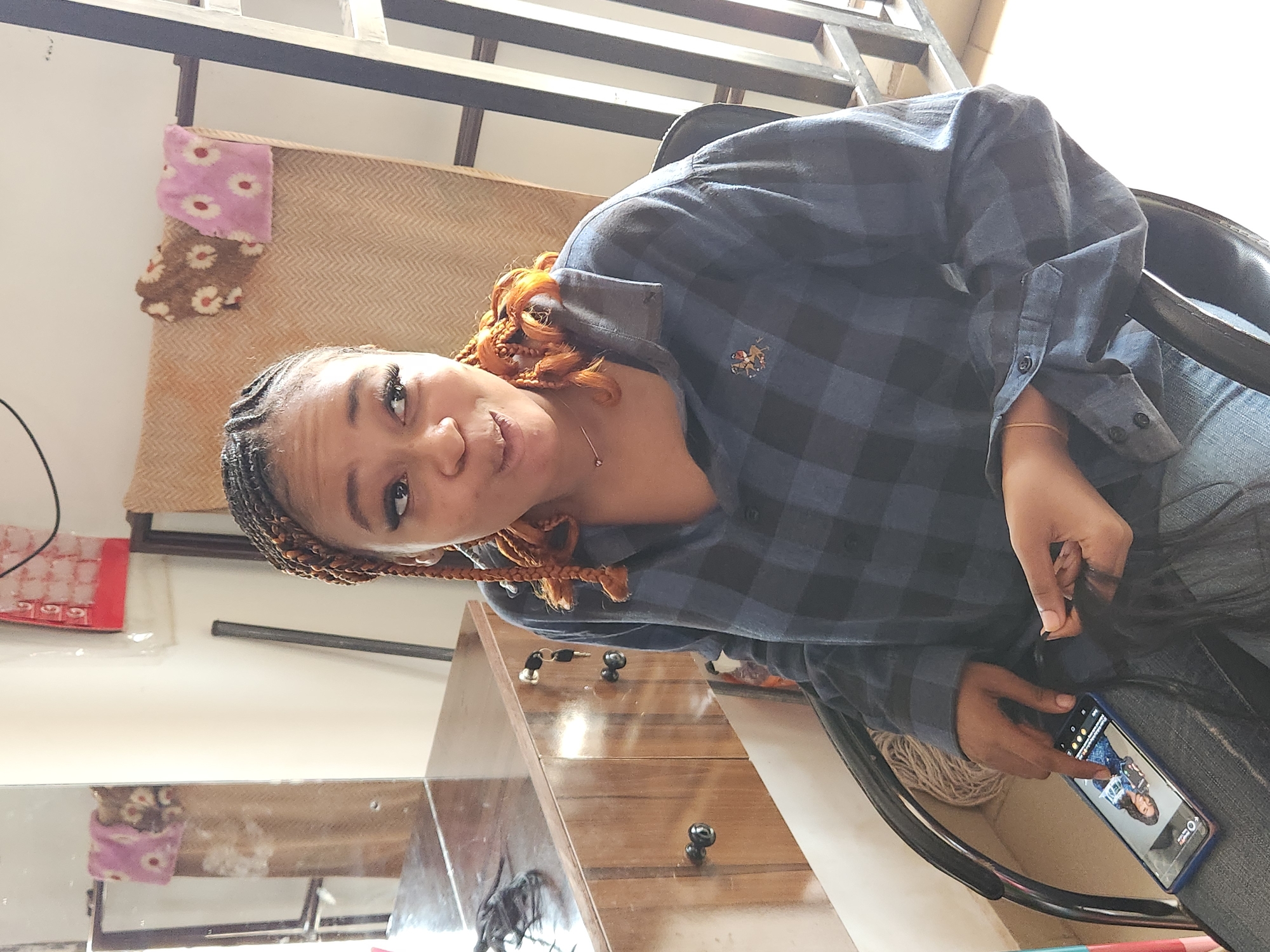 The image depicts a young Black woman with braided hair styled in two pigtails, adorned with copper-colored extensions. She is seated in a black chair, wearing a dark blue and black plaid button-down shirt and blue jeans. Her left hand is holding a black smartphone, displaying a picture of herself. Her right hand is engaged in braiding hair extensions. The background suggests an indoor setting, possibly a salon or a room with furniture. To the left, a wooden vanity with drawers and mirrors is visible, reflecting a pink and white towel hanging on a rack. A ladder-like structure stands to the right. The lighting indicates daytime. The location is identified as Sabon Tasha, Nigeria.