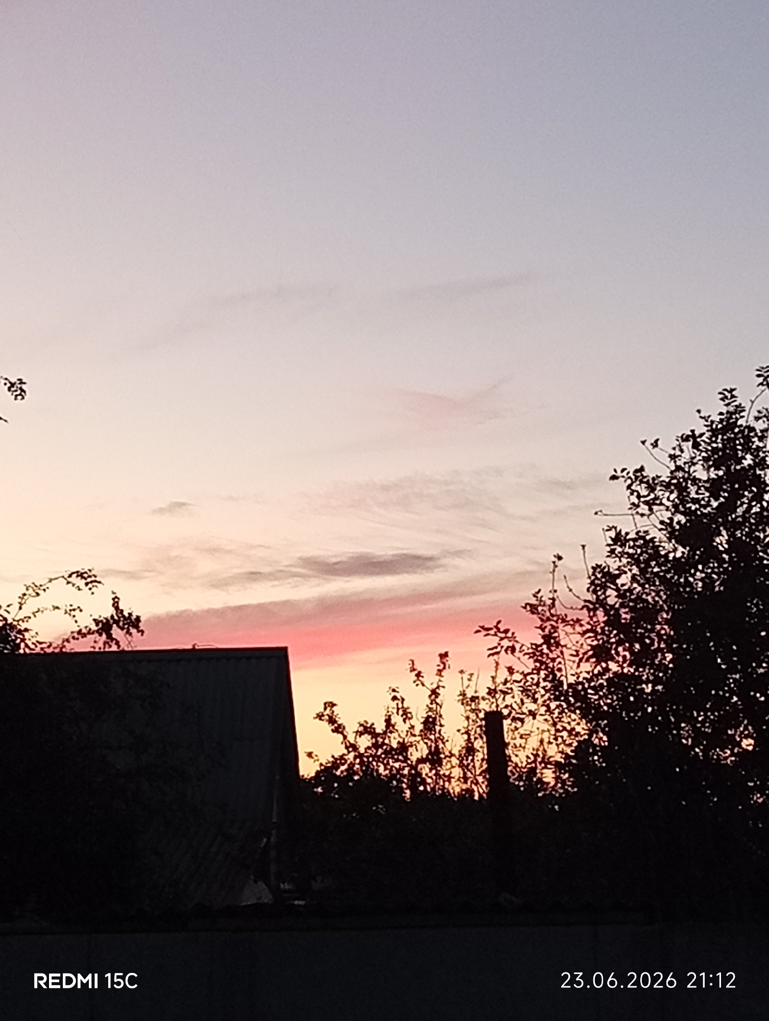 The image depicts a twilight scene with a pink and orange gradient sky. In the foreground, a dark silhouette of a house's roof is visible on the left, with trees and foliage silhouetted against the sky on the right. The bottom left corner displays "REDMI 15C" and the bottom right corner shows "23.06.2026 21:12". The location of Mykolaivka, Ukraine, is not discernible from the visual content of the image.