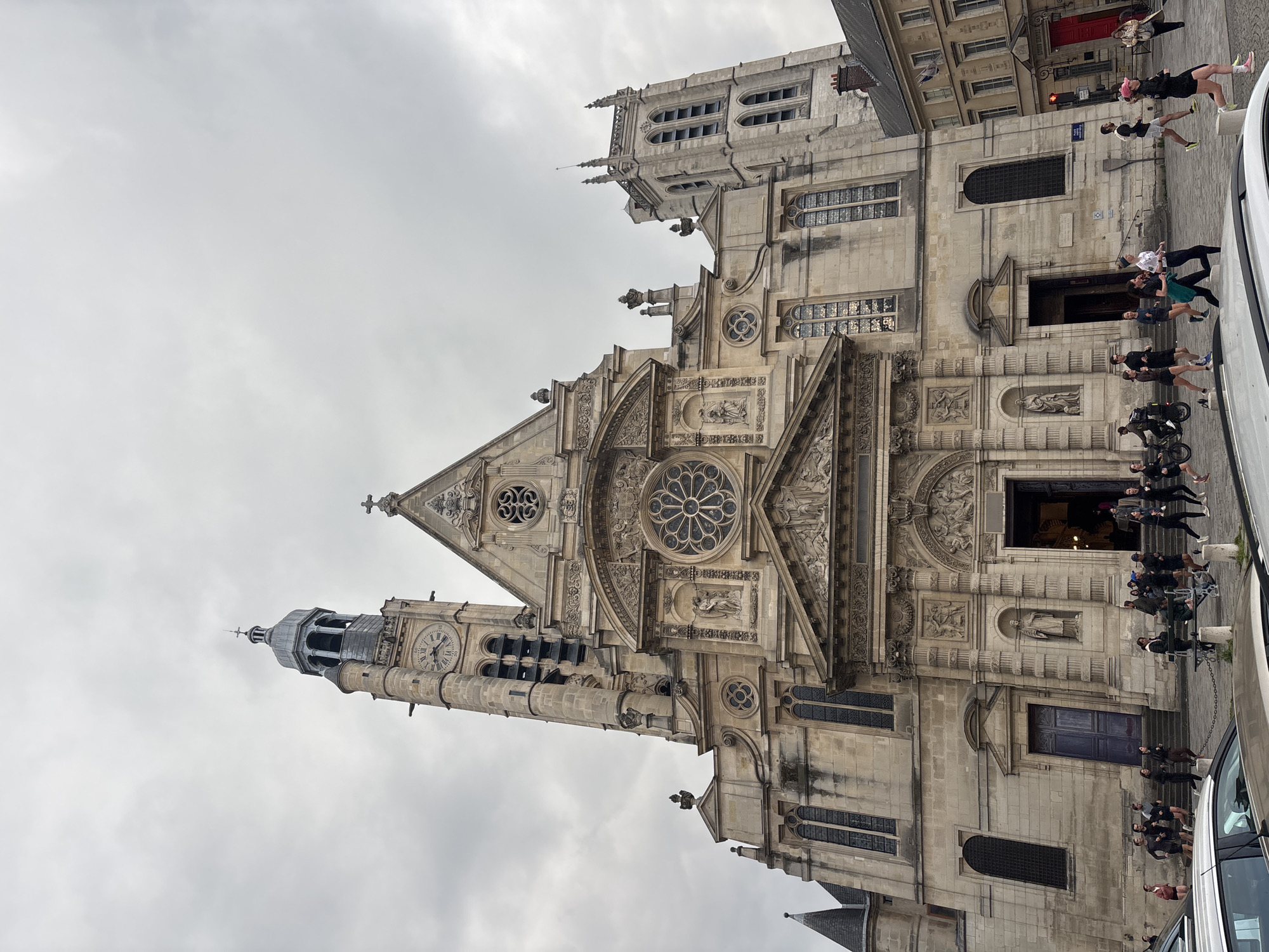 The image shows the Church of Saint-Étienne-du-Mont, a church in Paris, France, with a notable blend of Gothic and Renaissance styles. A large group of people is standing or sitting to the right of the frame in front of the church's facade. A vehicle is partially visible on the right side. The sky is overcast. The architectural details of the church are prominent, including its rose window, carvings, and clock tower. A street with buildings is also visible adjacent to the church.