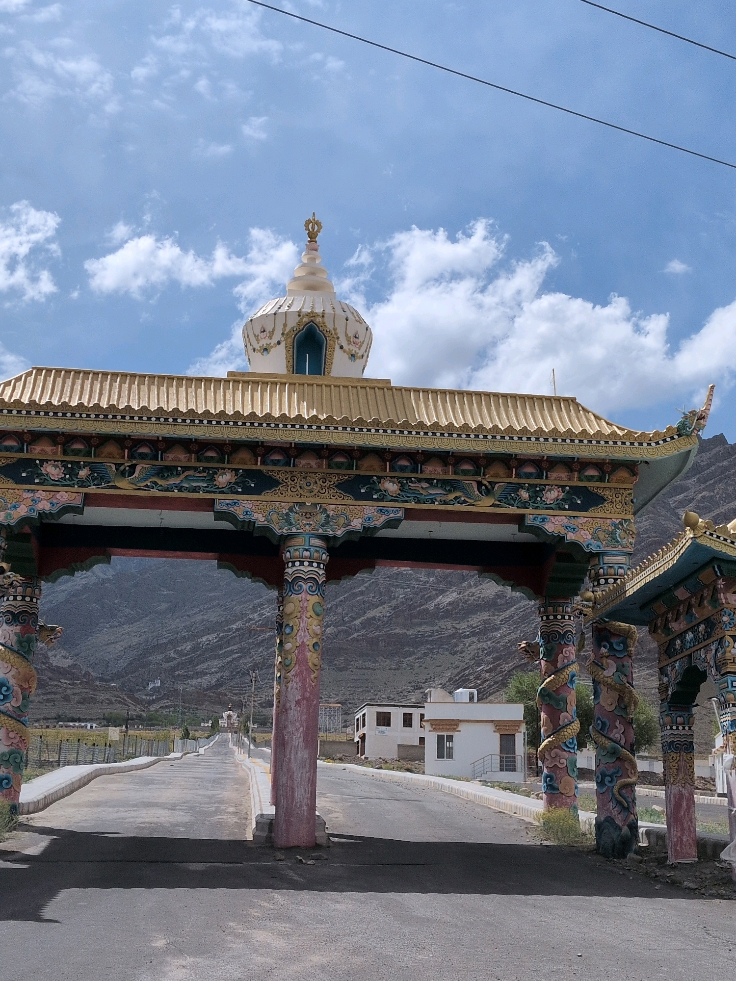 The image shows a ceremonial gateway in Martselang, India, with a stupa visible in the distance. The gateway is ornately decorated with colorful Buddhist motifs, including dragons and floral patterns, on its pillars and roof. The gateway is constructed with traditional Tibetan architectural elements, featuring a multi-tiered roof and a prominent stupa crowning the structure.
A paved road leads through the gateway, disappearing into the distance towards the mountains. The road is flanked by low walls and vegetation. In the middle ground, several white buildings with dark roofs are visible, typical of the region's architecture. The sky is blue with scattered white clouds, and the sun appears to be shining, casting shadows on the road.
The surrounding landscape is mountainous and arid, with dry, rocky slopes visible behind the gateway and buildings. The overall scene suggests a serene and spiritual atmosphere, common in Buddhist pilgrimage sites. No people are visible in the image.