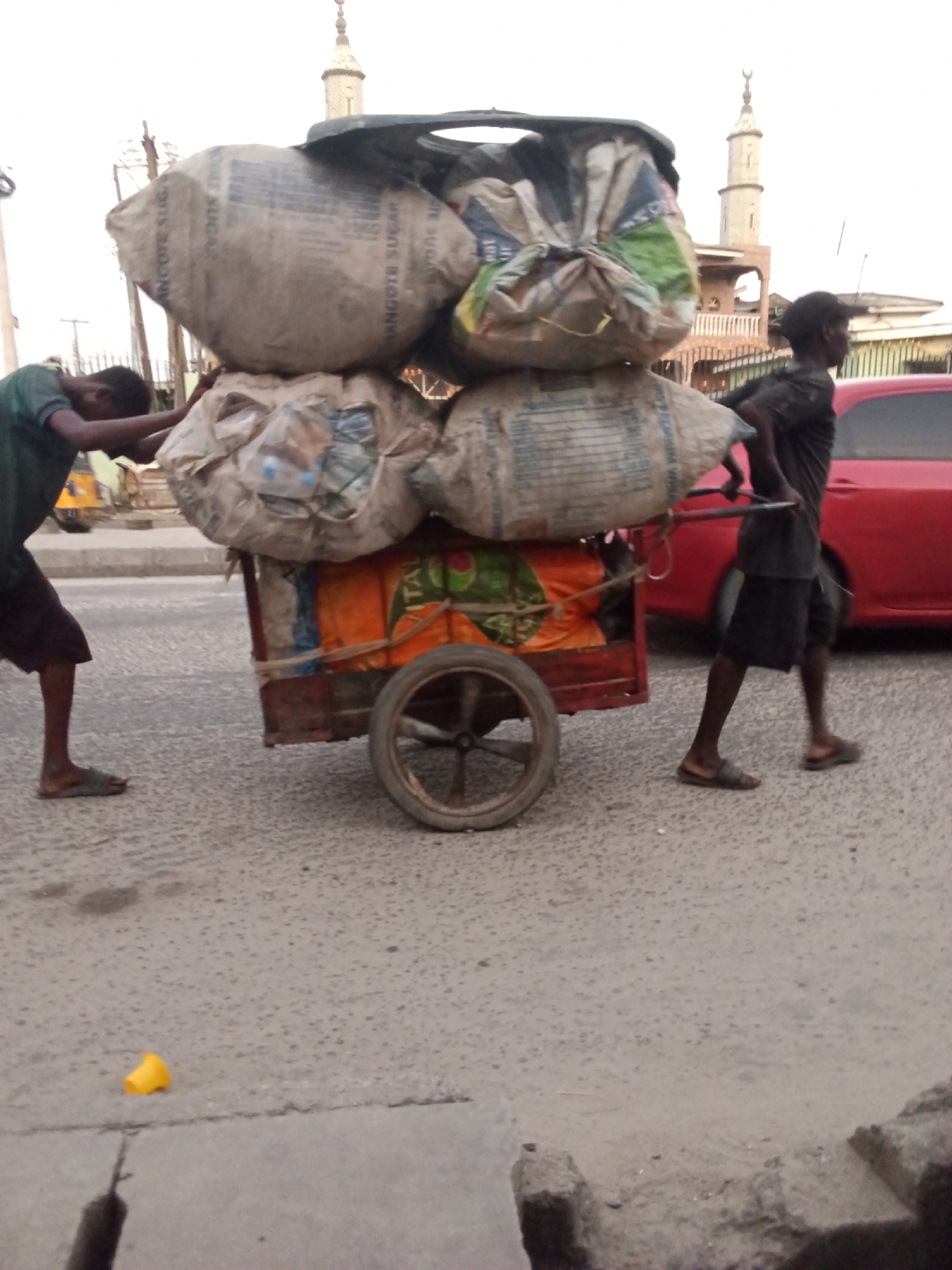 This image captures two men manually transporting a heavily loaded handcart on a paved road in Ado, Nigeria. The diffused, bright lighting suggests either early morning or late afternoon.
On the left, a man in a dark green t-shirt and shorts, wearing flip-flops, pushes the cart from behind, his body leaning into the effort. On the right, another man, also in a dark t-shirt and shorts with flip-flops, pulls the cart by its handles, walking forward.
The two-wheeled handcart is laden with a towering stack of large, bulging sacks, which appear to contain light, bulky items like plastic waste or recyclables. Visible text on some of the sacks clearly reads "DANGOTE SUGAR" and "DANGOTE CEMENT," indicating the reuse of industrial bags. An orange tarp, partially visible on the cart, also shows the faint word "VITAL."
In the background, a red car is parked on the right side of the road. Prominently rising above distant buildings is a tall, light-colored minaret topped with a crescent moon, signifying a mosque. Utility poles and wires are faintly visible on the left. The road surface is asphalt or concrete, showing some wear, and a small yellow object lies on the ground in the foreground. The scene depicts strenuous manual labor in an urban or semi-urban environment.