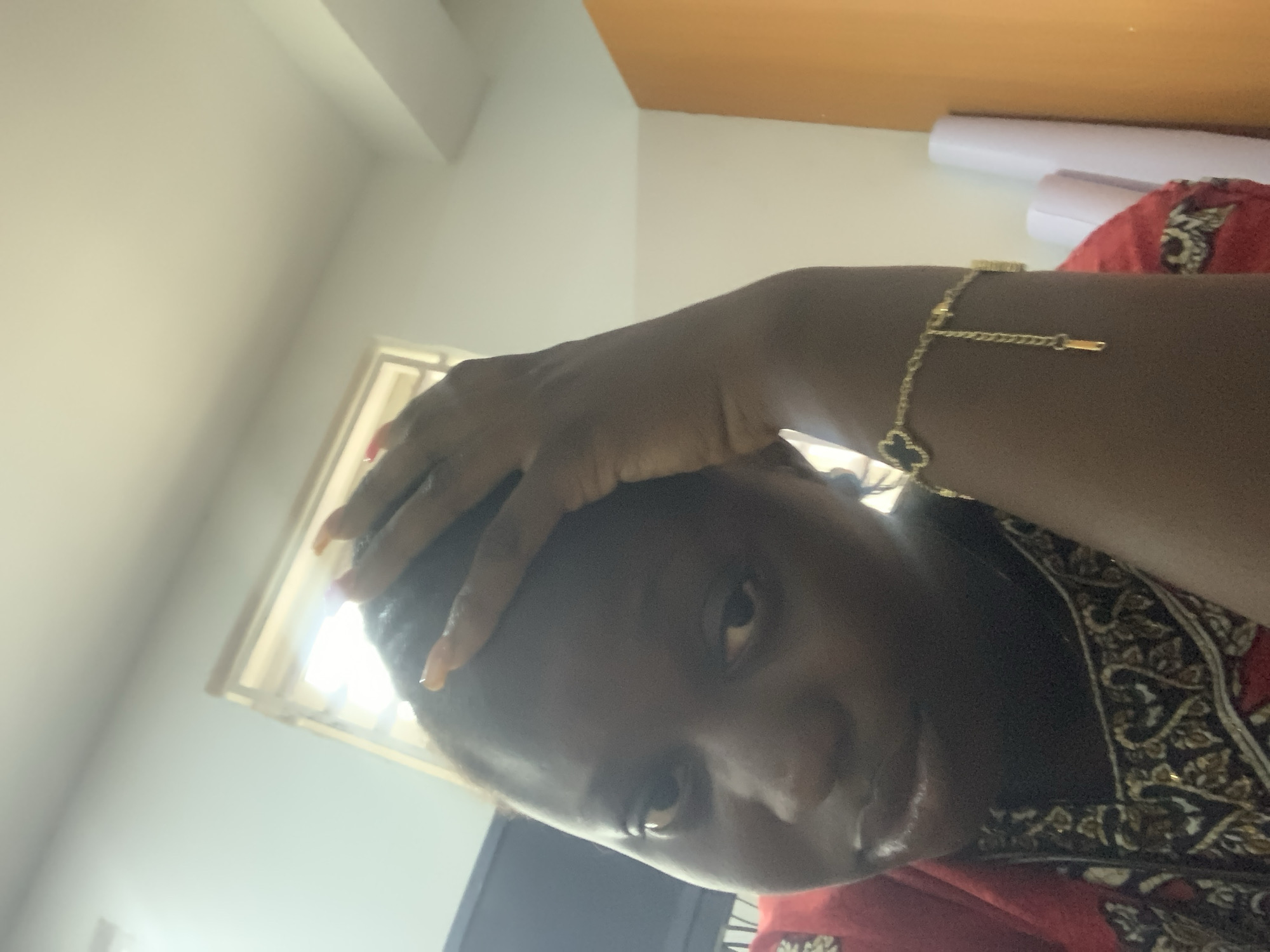 A dark-skinned woman is pictured from the chest up, with her right hand on her head. She is wearing a red and black patterned garment and a gold bracelet on her right wrist. The background shows a window with natural light, a portion of an orange wall, and two rolled-up white papers. The image appears to be taken indoors.