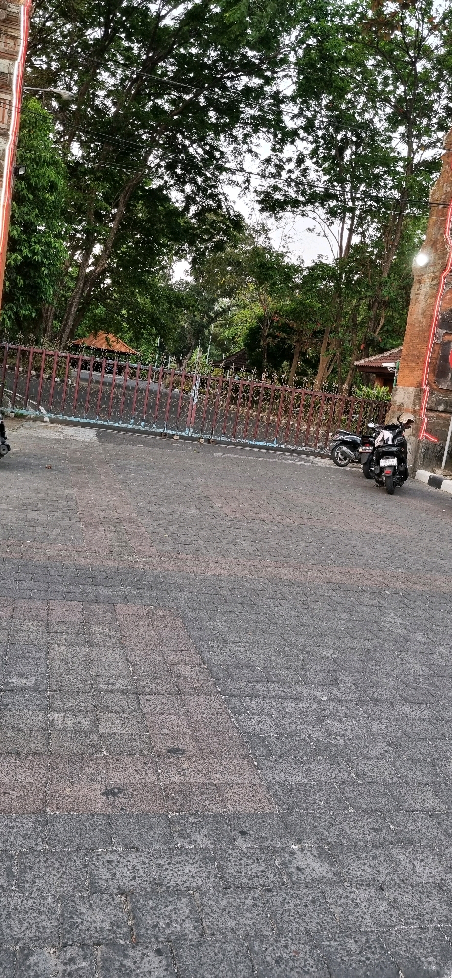 An outdoor scene features a large metal gate with intricate scrollwork, a brick pillar adorned with red neon lighting, and two parked motorbikes. The ground is paved with dark grey and red interlocking stones. Beyond the gate, trees and the roof of a small structure are visible. The lighting suggests it is either dawn or dusk. The location is an entrance to a property in Denpasar, Indonesia.