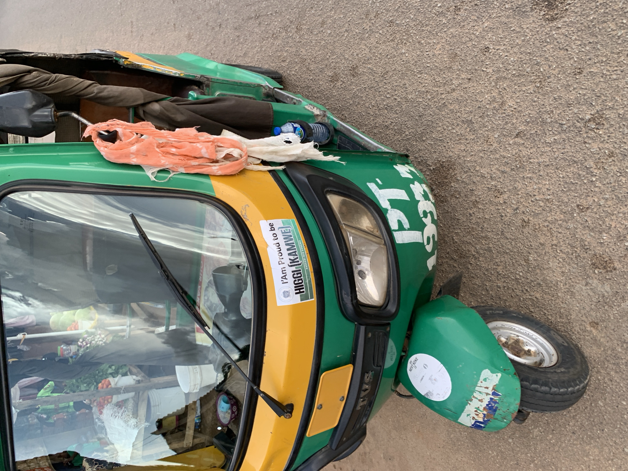 The image shows the front and side of a green and yellow auto-rickshaw parked on a dirt road. The auto-rickshaw has a yellow bumper on the front, and a black grille with the "TVS" logo. There is a windshield with windshield wipers. A sticker on the windshield says "I'm Proud to be HIGGI | KAMWE". A piece of orange and white cloth is hanging from the side mirror. A bottle of water is in a holder on the side. A circular sticker on the front fender says "MY NEXT KEKE IS..." The auto-rickshaw is a mode of transport common in Jos, Nigeria. The scene suggests it is a street scene in Jos, Nigeria.