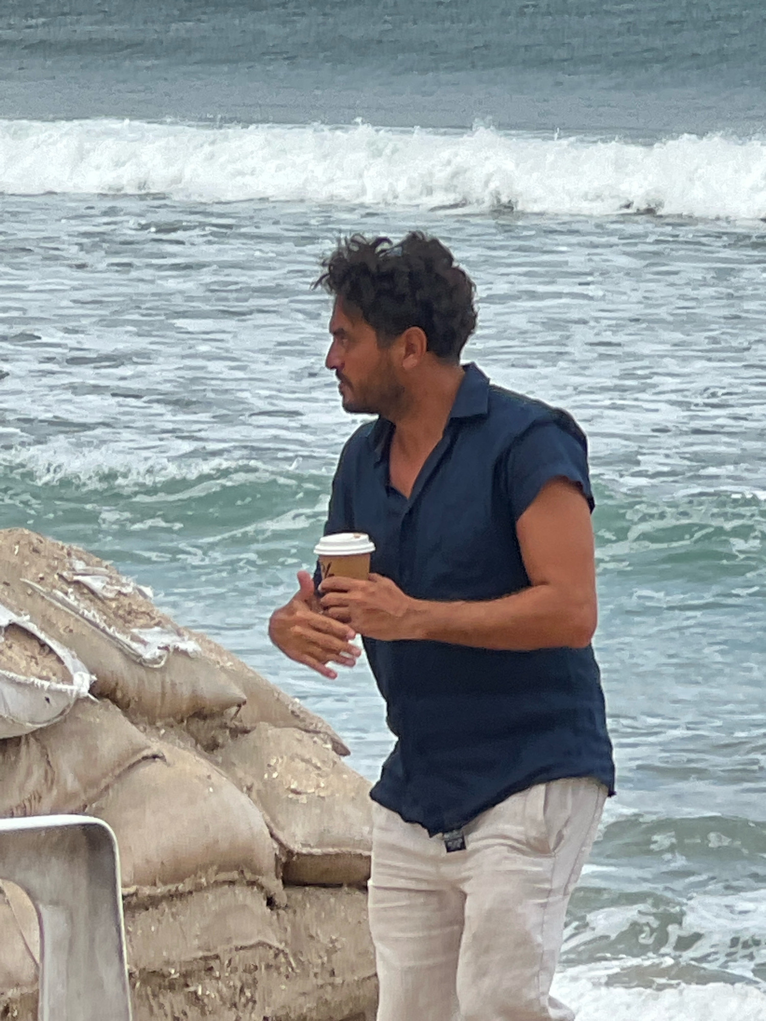 A man with dark, curly hair stands on a beach in Kuta, Indonesia, holding a disposable coffee cup. He is wearing a dark blue short-sleeved collared shirt and light-colored pants. The man is looking to his left, with waves and the ocean in the background. In the foreground to the left of the man is a pile of sandbags.