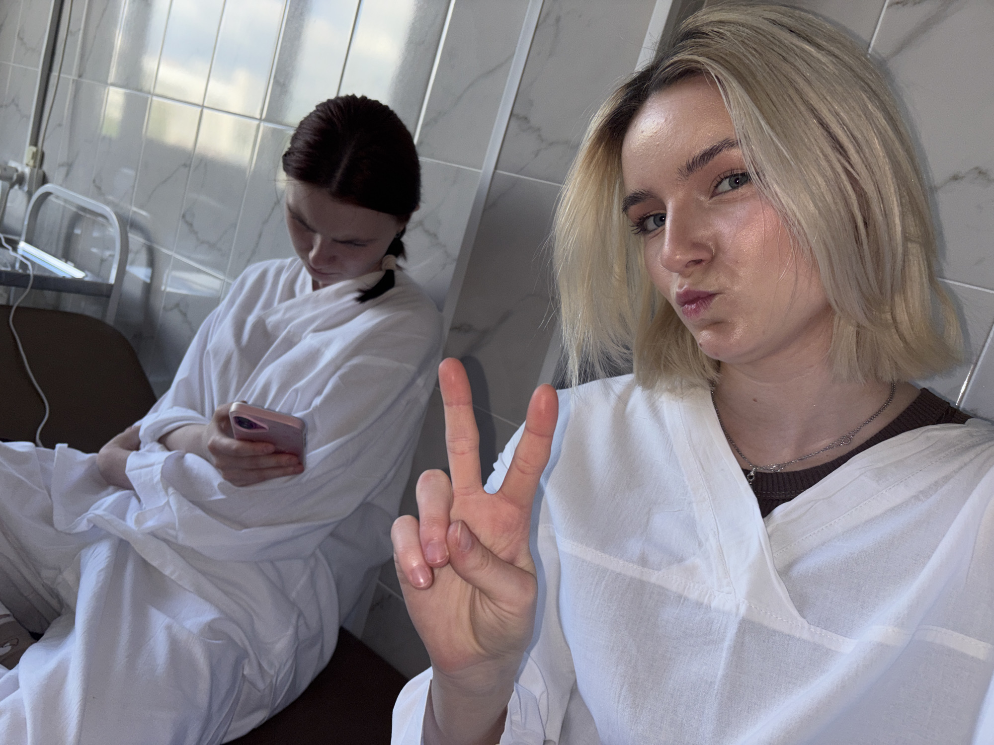 This media file captures two young women indoors, likely in Saint Petersburg, Russia, against a backdrop of white tiled walls, suggesting a spa, bathhouse, or medical facility.
In the foreground, a young woman with short blonde hair and blue eyes is taking a selfie. She wears a white robe over a dark brown top and a silver chain necklace. Her lips are playfully pouted, and she makes a peace sign with her right hand, conveying a relaxed and cheerful demeanor.
Behind her, seated to the left, is another woman with dark hair tied back, also dressed in a white robe. She is focused on a pink smartphone held in her hands, looking downwards and appearing engrossed in its contents.
The room features light-colored, possibly marble-patterned, tiled walls. A metal railing is partially visible in the upper left corner of the background, and some brown furniture, possibly a chair or bed, is also present. The lighting is soft and even, indicating an indoor setting without direct sunlight. No visible text is present in the image.