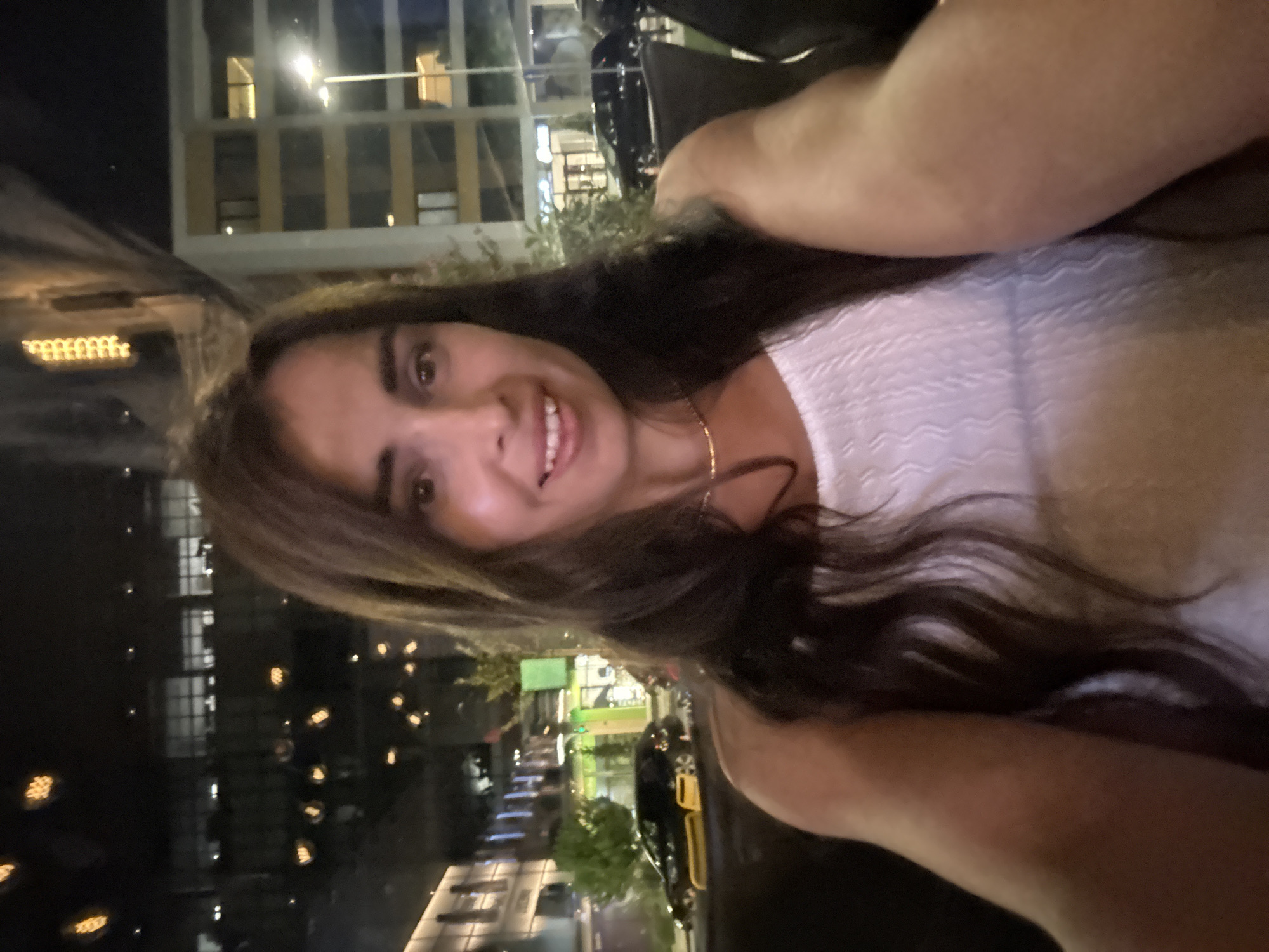 A woman with long, dark hair and fair skin smiles at the camera in a selfie. She is wearing a white, sleeveless top and a delicate gold necklace. The background suggests an urban setting at night, with buildings, streetlights, and parked cars visible. The lights from the buildings and street are reflected in the glass in front of the photographer.