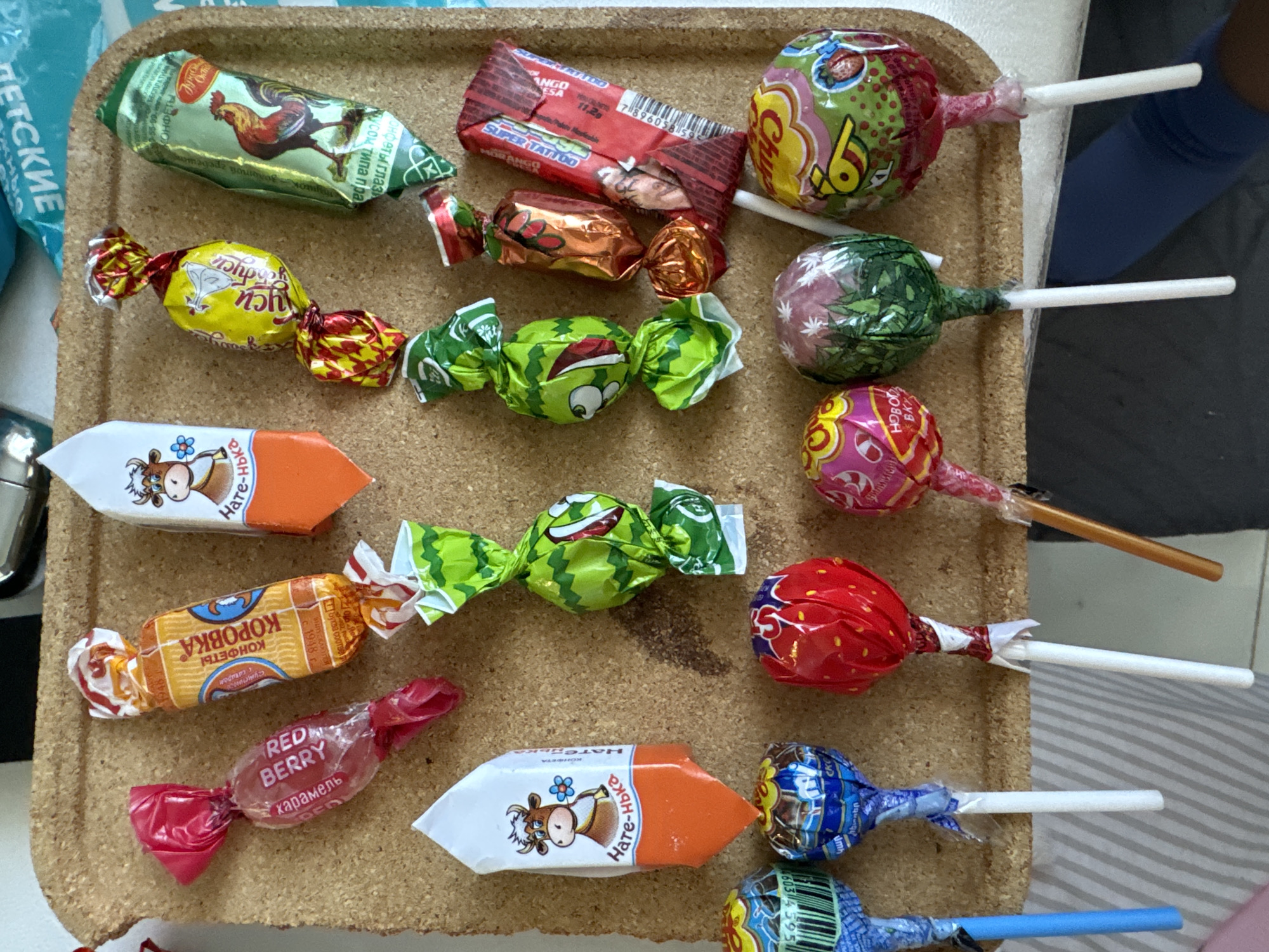 A collection of candies, including lollipops and wrapped hard candies, sits on a cork-textured tray. The candies are of various colors and packaging. Lollipop brands are Chupa Chups. Wrapped candies include "Red Berry", "Korovka", "Super Tattoo" mango flavor, and "Natenka". The candies and tray are resting on a light-colored striped surface, and a person's legs are visible in the background. The location in the city of Akulovo (Gp Kubinka), Russia is not discernible from the image.