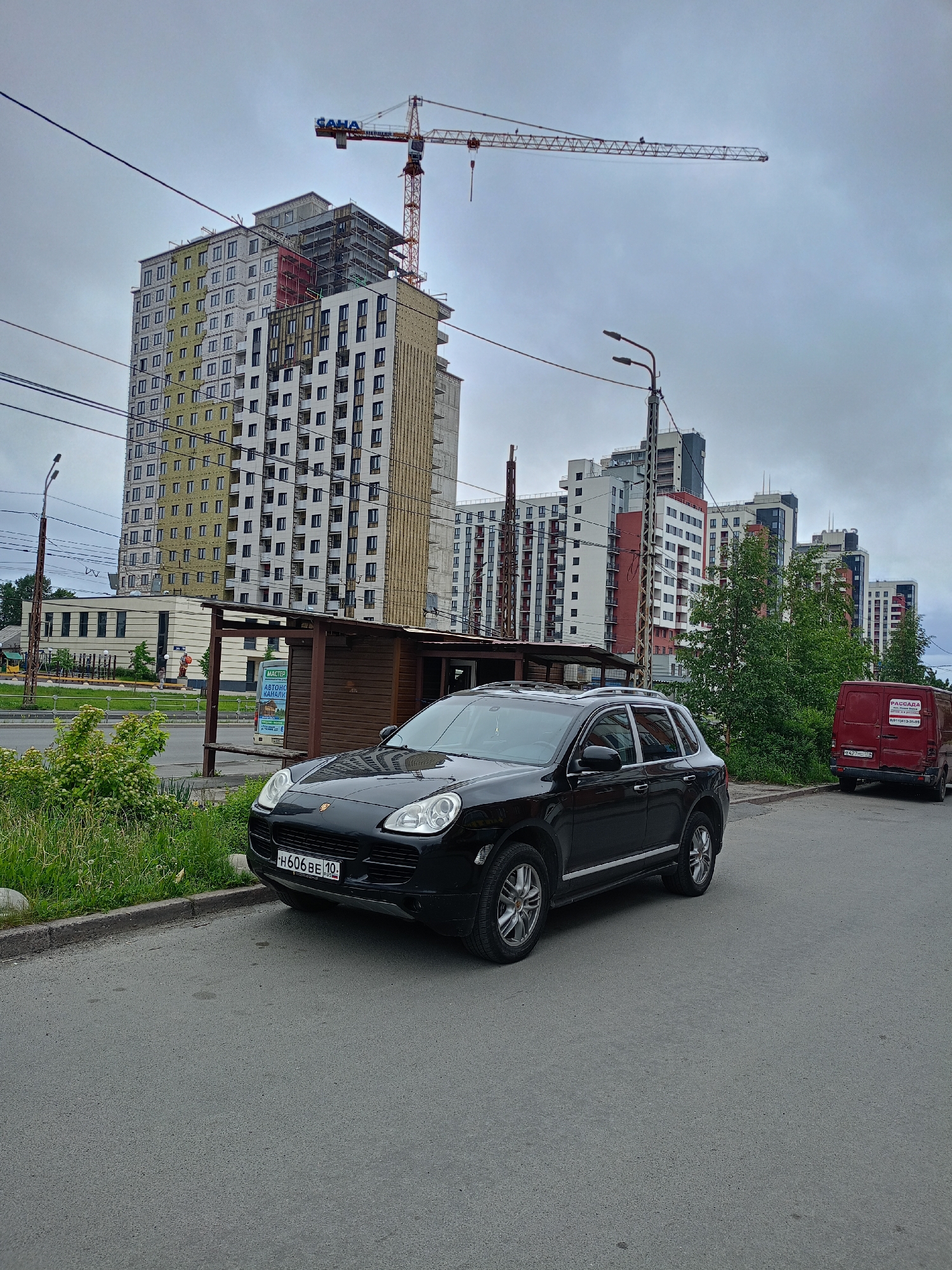 A black Porsche Cayenne is parked on an asphalt road in front of a bus stop. The bus stop is made of wood and has a sign that reads "MACTER ABTONOGOCM KANOM". In the background, there are several tall apartment buildings under construction, with a large construction crane overhead. A red van is parked behind the Porsche. The sky is overcast.