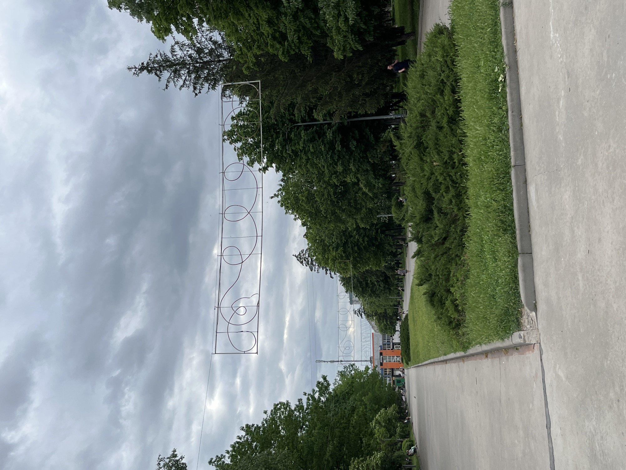 The image shows a street in Kryvyi Rih, Ukraine, on a cloudy day. The road is concrete and has a low concrete border where the pavement ends. On the right side of the road there is a green area, then trees. Trees line both sides of the road. There are decorative metal signs with circles that are suspended above the road. Some buildings can be seen between the trees in the distance. A person in dark clothing can be seen lying on the grass next to the trees. Other pedestrians are also visible along the street.