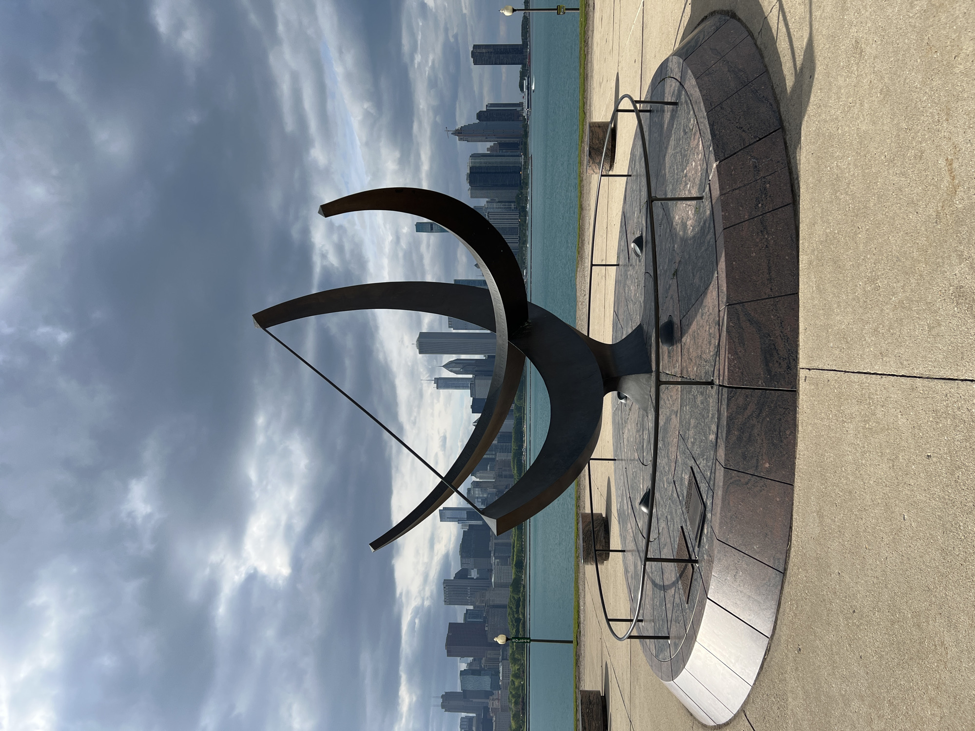 The image shows a large sundial sculpture located along the lakefront in Chicago, Illinois, United States. The sculpture is composed of dark metal arcs and a dial made of dark stone tiles, positioned on a concrete surface. In the background, the Chicago skyline is visible across the lake, under a sky with dramatic clouds. The overall perspective is a slightly tilted, ground-level shot.