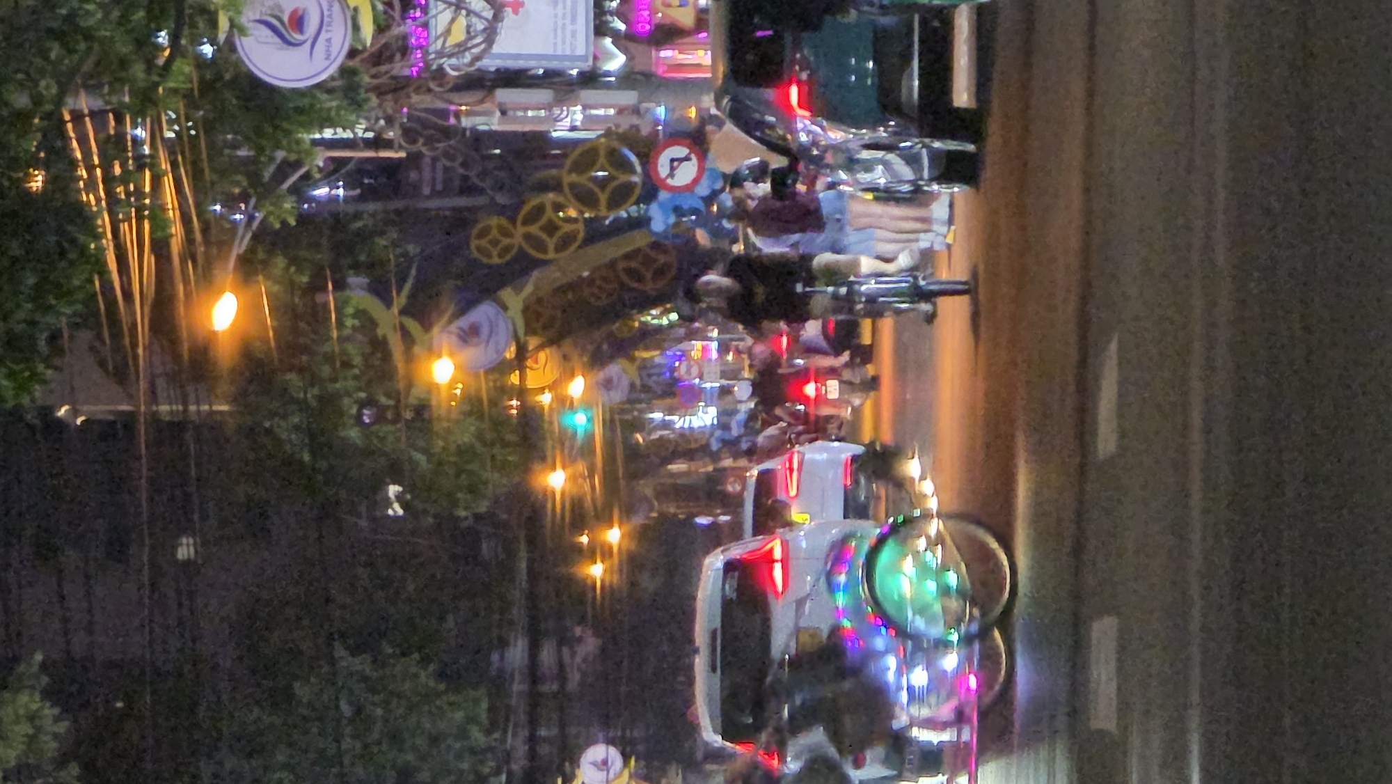 This media file captures a vibrant street scene at night in Nha Trang, Vietnam. The image is oriented vertically, showing a busy urban environment illuminated by numerous artificial lights.
**Subjects and Activity:**
The street is populated with various vehicles, including multiple motorbikes and at least one white car, suggesting active evening traffic. Several individuals are visible, primarily on motorbikes, either riding or stopped. One person is clearly seen operating a motorbike with a passenger behind them, while another person is seated on a separate motorbike further back. The scene depicts everyday commuting or travel activity during nighttime.
**Notable Details:**
The time of day is clearly night, evidenced by the dark sky and the prominence of artificial light sources. Streetlights cast warm orange glows, while vehicle headlights and taillights add to the illumination. A green traffic light is visible, as are several red taillights from cars and motorbikes.
The street is adorned with festive or decorative lighting and structures. An archway-like structure features prominent golden gear-shaped decorations, along with other abstract shapes. A large round white sign is visible at the top left, clearly displaying the text "NHA TRANG" and a logo, definitively identifying the location. Another red circular road sign with a white bar and an arrow pointing right with a line through it indicates a "no right turn" restriction.
The overall atmosphere is lively and colorful due to the diverse light sources and decorations.
**Visible Text:**
The most prominent visible text is "NHA TRANG" on the white circular sign.