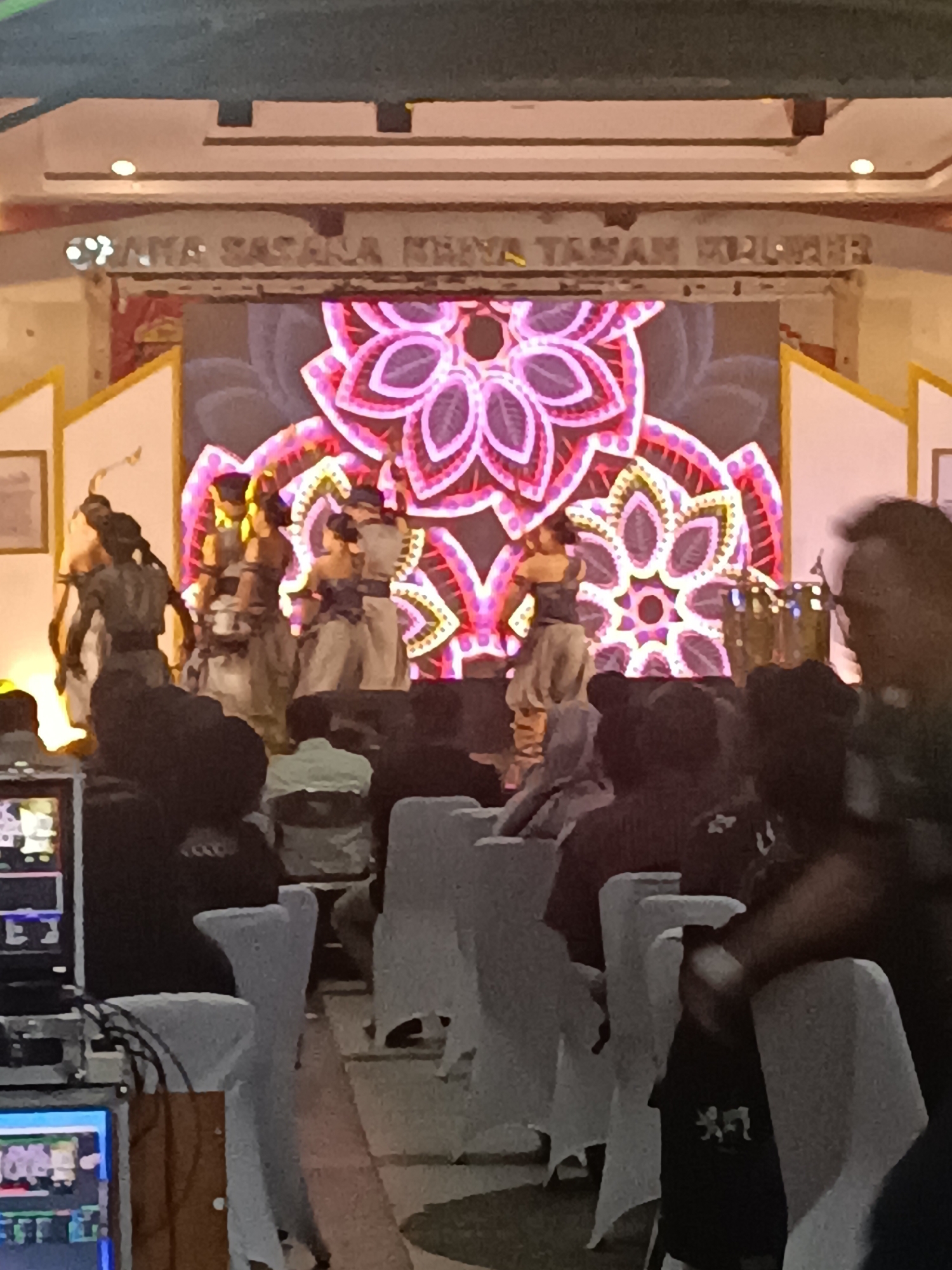 The image depicts a performance taking place on a stage decorated with a large, vibrant floral pattern projected onto a screen. Several dancers, dressed in traditional-looking attire, are performing on stage. In the foreground, an audience is seated in rows of white chairs, with some individuals visible in the lower half of the image. To the left, there is a sound and lighting console with monitors displaying various information. The scene suggests an event or cultural performance, likely in a public venue within Condongcatur, Indonesia, given the context provided by the prompt, although no specific landmarks confirm this location within the image itself.