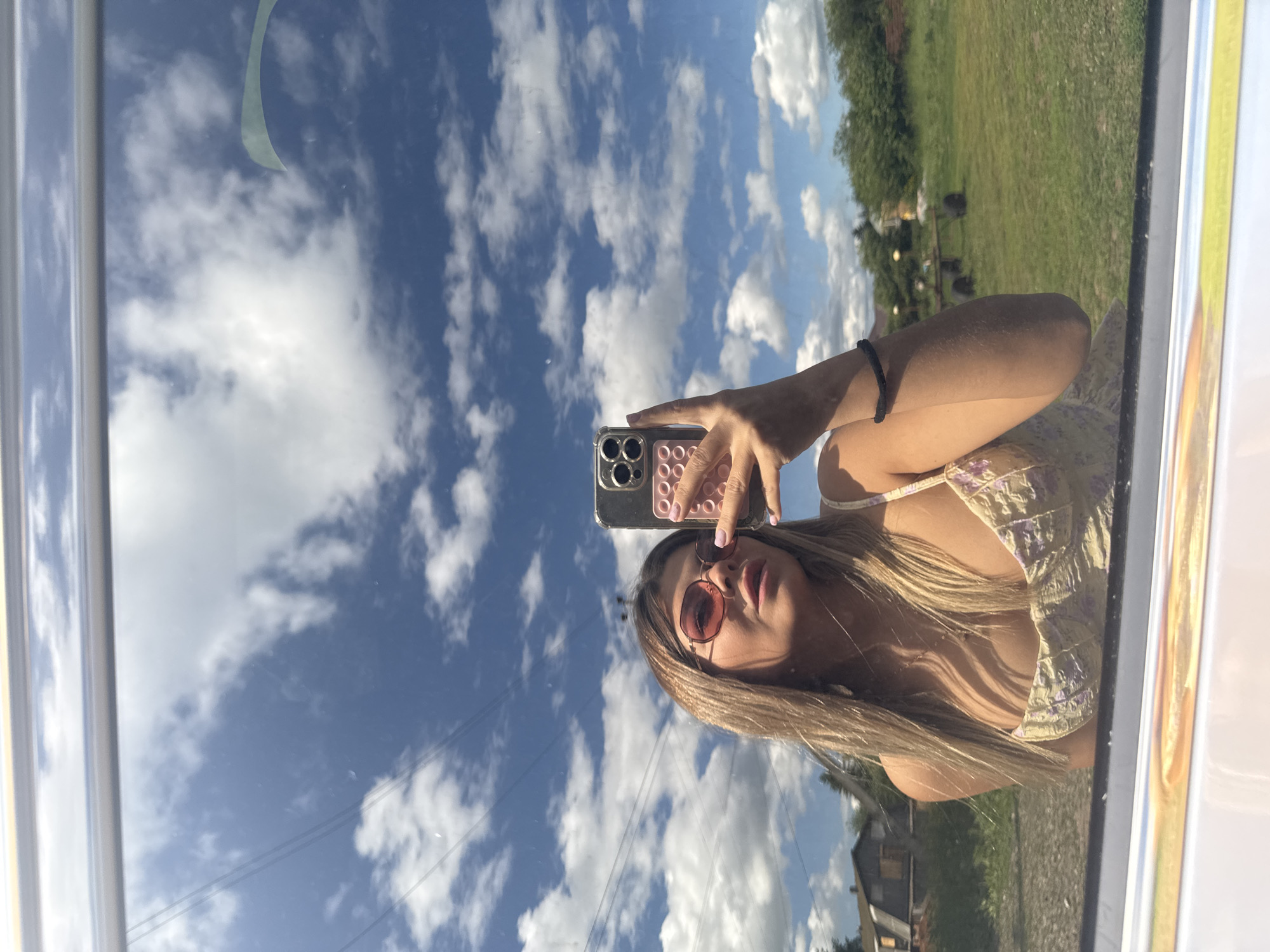A young woman with blonde hair and sunglasses is taking a selfie in a reflective surface, possibly a window or car door, which is framing the lower part of the image. She is wearing a light-colored floral top. The reflection captures a bright, sunny day with a blue sky filled with white, fluffy clouds. In the background of the reflection, there is a rural landscape with some buildings, a grassy field, and trees. The image is shot from a slightly low angle, looking up at the woman and the sky. There is no discernible indication of the city of Pastukhovo, Russia in the image.
