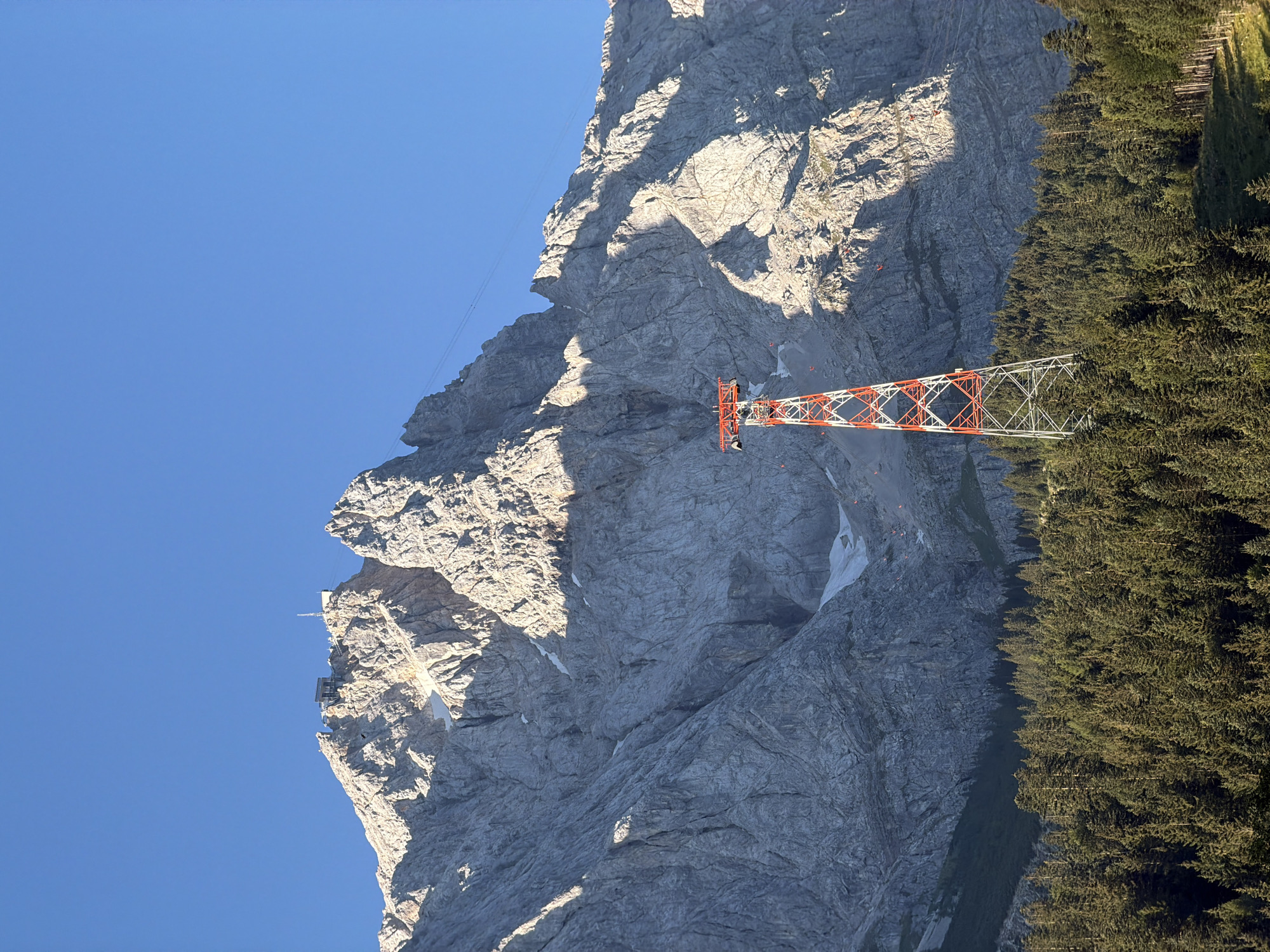 This is a vertical, medium shot of a ski lift tower in front of a large mountain range under a clear blue sky. The ski lift tower is red and white with a lattice structure, and the cable extends upwards towards the mountain. The mountain is steep and rocky, with some patches of snow and vegetation. The base of the mountain is covered in a dense forest of evergreen trees. The sun is shining, casting shadows on the mountain. The image appears to be taken from a distance, with the ski lift tower prominently featured in the foreground. This is likely the Zugspitze, Germany's highest peak, located near Grainau.