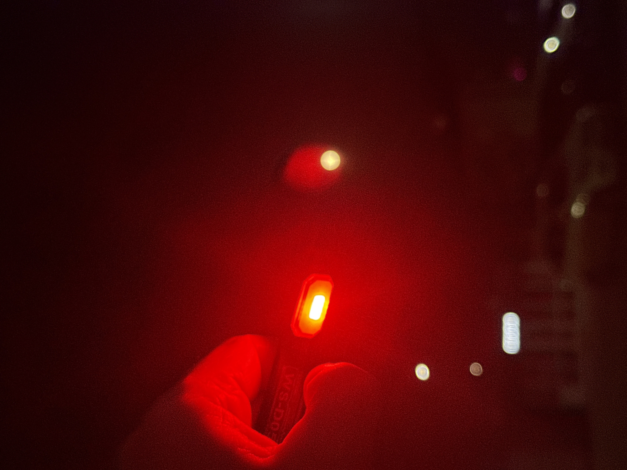 A person's hand holds a small, rectangular device with a glowing white light in a red-lit environment. The device has markings, possibly text, on its side. The background is blurred and features indistinct light sources. The location or information of the city of Votkinsk, Russia is not present in the image.