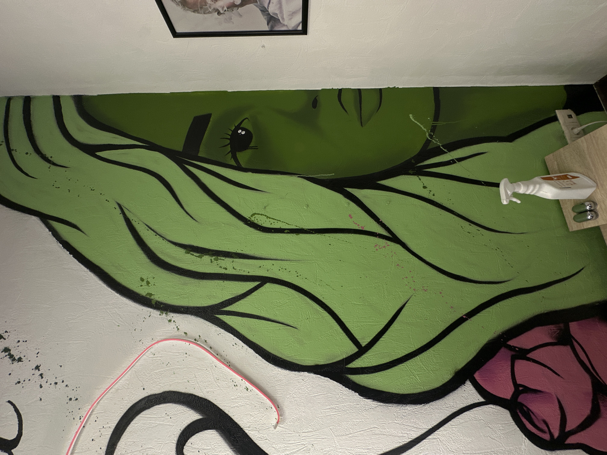 The scene depicts an interior wall in Saratov, Russia, dominated by a large, stylized mural. The mural on the left features a woman's profile, rendered with light green skin and hair, outlined in black. Darker green shading is visible within the hair strands. A partial view of a pink or magenta element, possibly a rose, appears at the bottom left of the mural. The underlying wall surface is textured and off-white. Numerous small green and pink paint splatters are scattered across both the white and green sections of the wall. A thin, wavy pink cable is attached to the upper left white wall section.
Below the mural, positioned on a light-colored wooden surface, are a white plastic spray bottle with an orange label and two small, silver-colored cylindrical objects. An electrical outlet with a white cord plugged into it is visible on the wall beneath this surface. To the right, on an adjacent plain white wall, a black-framed picture shows the upper body of a person, likely male, with visible smoke or vapor emanating from their mouth. No individuals are present in the immediate scene.