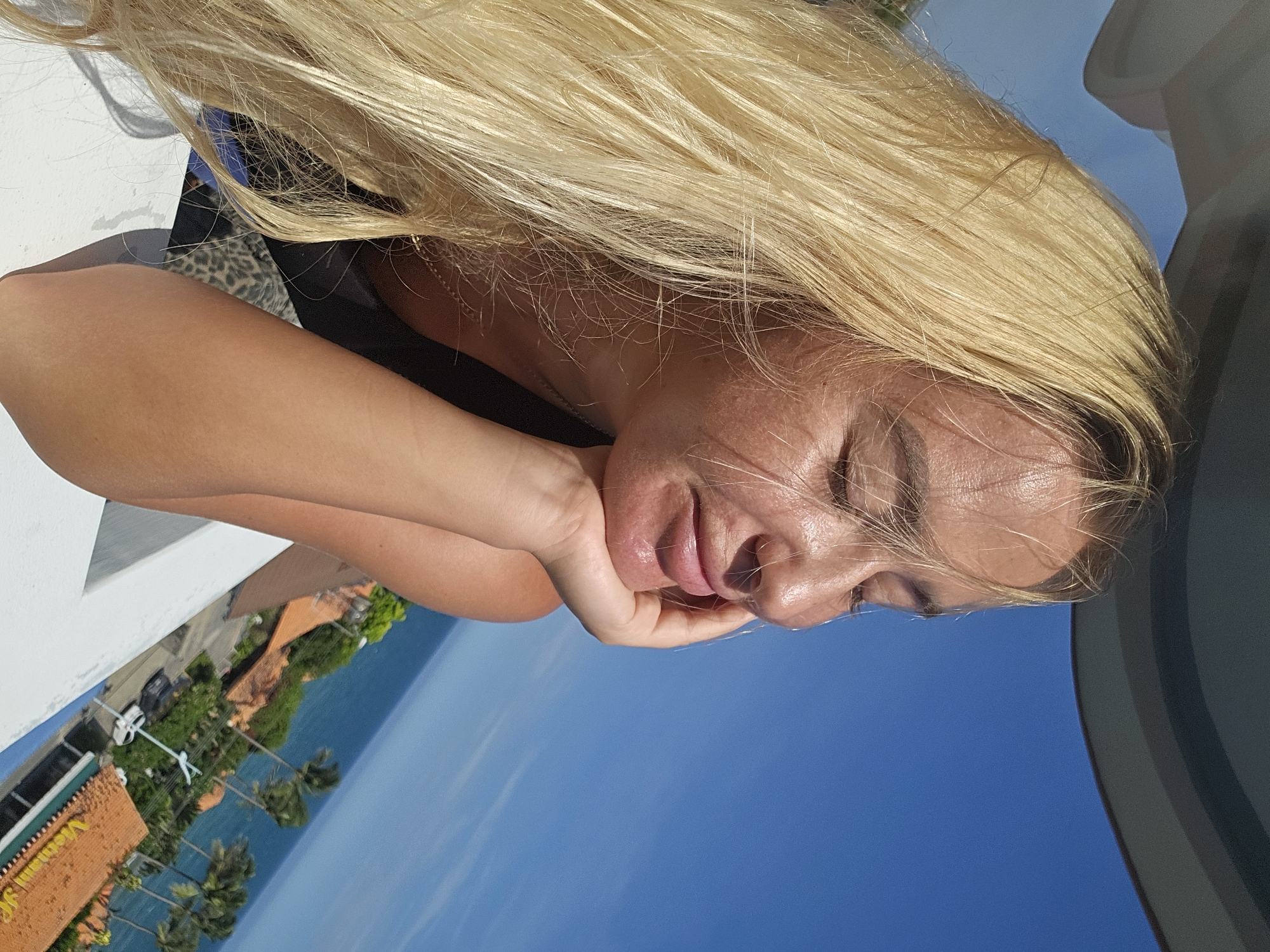 A blonde woman with her eyes closed is resting her chin on her hand. She is looking out at a body of water and palm trees. The background shows a coastal town with red-tiled roofs and a blue sky. The woman is wearing a black tank top and a necklace. The image appears to be taken on a sunny day. The location is Phu Thuy Ward, Vietnam.