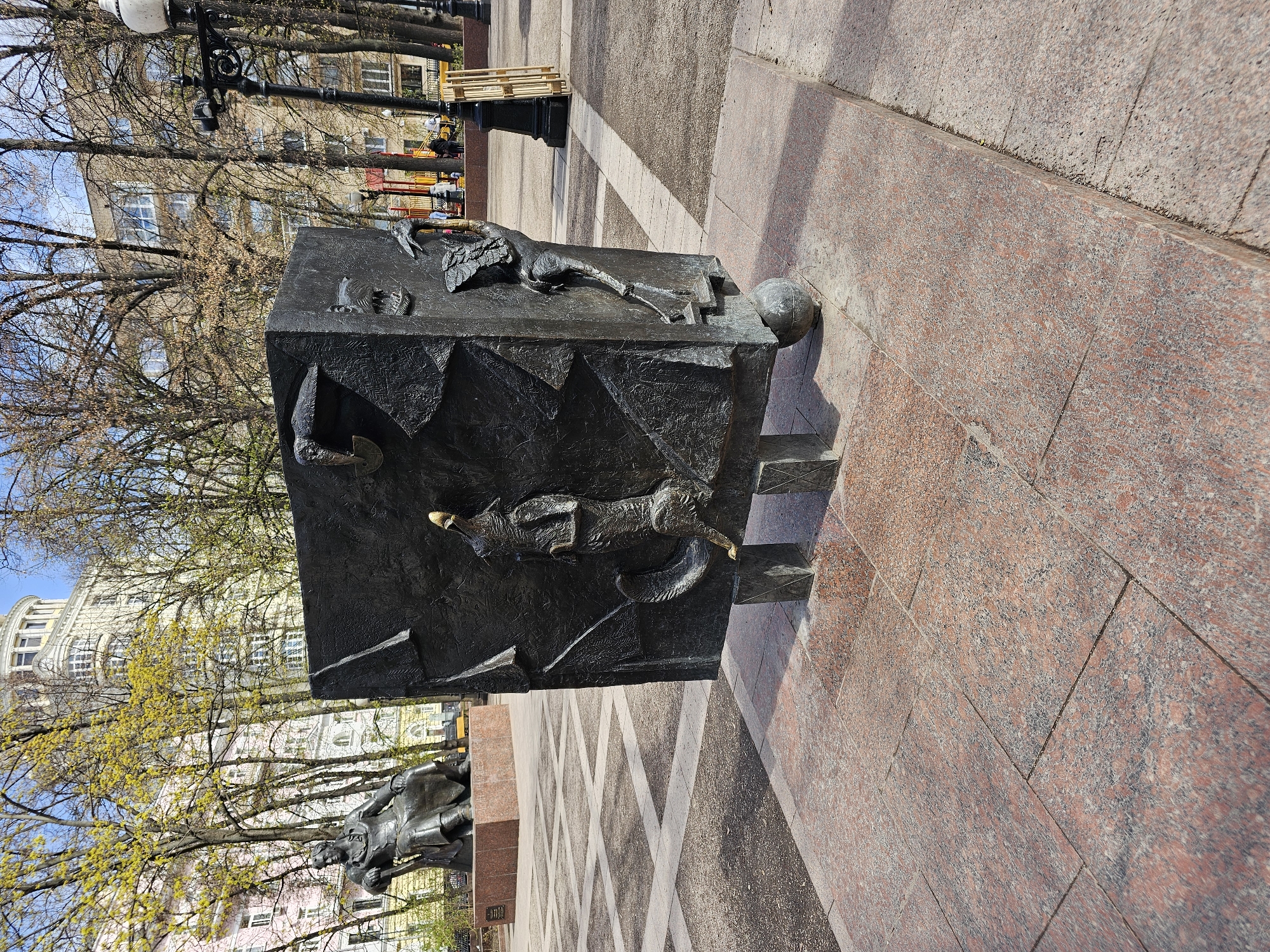 The image shows a bronze sculpture of Antoine de Saint-Exupéry’s “Little Prince” in Moscow, Russia. The sculpture is a cube-shaped monument depicting scenes from the book, with figures of the Little Prince, a fox, and other characters. The sculpture sits on a red granite pedestal and is located in a public square or park area. Buildings and trees are visible in the background, indicating an urban setting. The pavement around the sculpture is composed of patterned tiles.